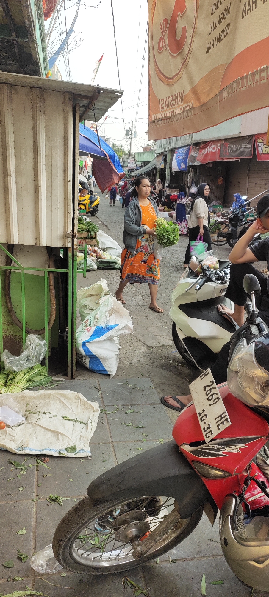 A woman in an orange dress with a grey jacket walks through a market in Karangtowo, Indonesia, holding a bunch of leafy green vegetables. She is walking on a tiled sidewalk, which is littered with fallen leaves and debris. To her left, a white and green stall is visible, with more vegetables displayed outside. Behind her, a yellow motorcycle is parked. Further down the street, other people are seen walking and browsing market stalls, with more motorcycles parked along the way. A large banner with text is hanging above the market stalls in the background. The scene depicts a bustling outdoor market environment.