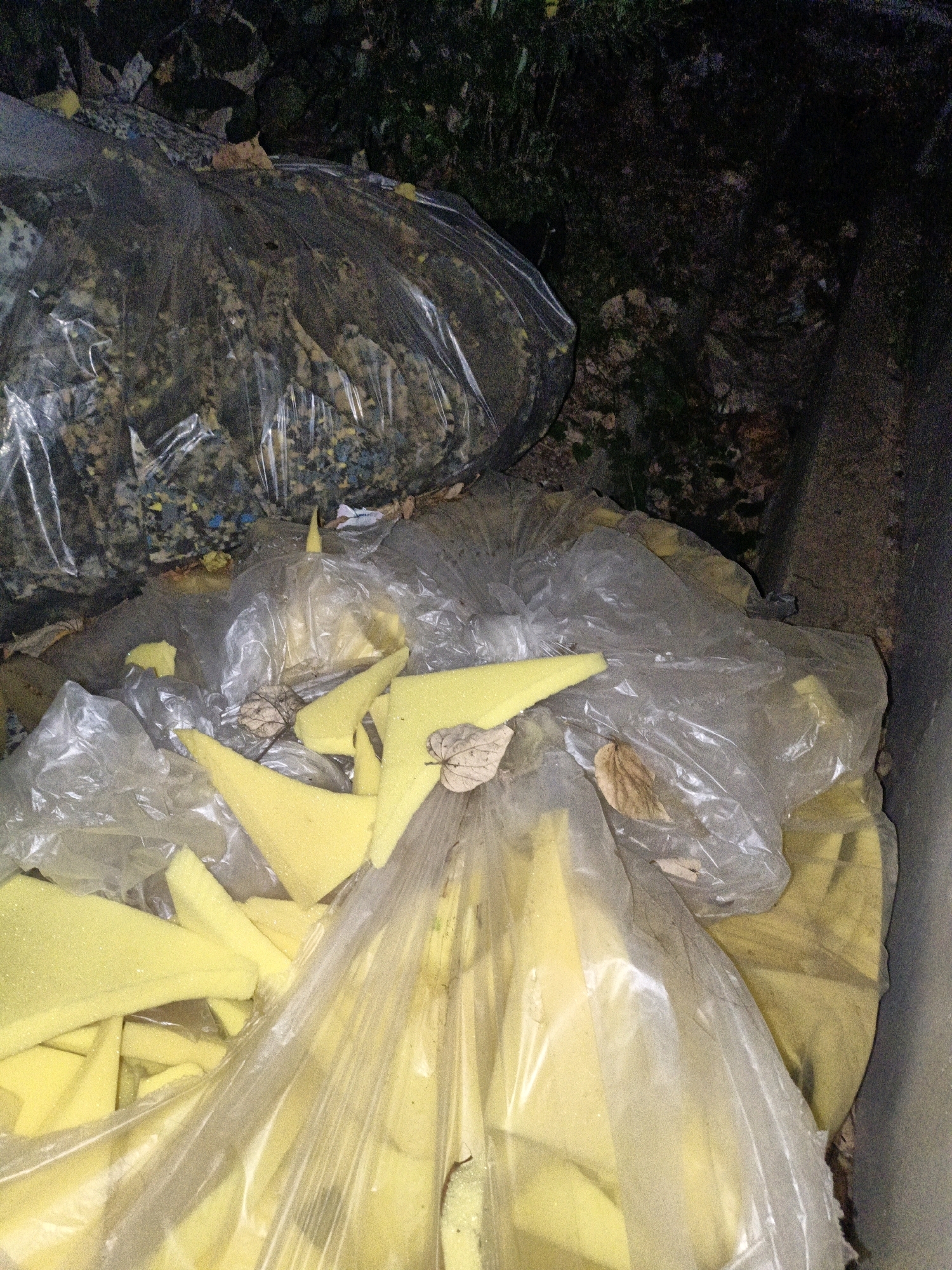 In Cikal, Indonesia, within a concrete structure that appears to be a receptacle or bin, lies a collection of discarded materials. The primary subjects are several large, clear plastic bags filled with yellow foam pieces of various sizes and shapes. Some pieces are triangular, others rectangular, suggesting they are remnants of cushions or padding. The bags are translucent, allowing the yellow foam to be the dominant visual element. In the background, a larger, opaque plastic bag is visible, filled with a mix of dark, granular material, possibly mixed with shredded foam or other debris. The environment surrounding the bin is dark and appears to be outdoors, with hints of foliage and shadowed ground. The lighting is dim, suggesting the image was taken at night or in a poorly lit area. No people are present in the scene.