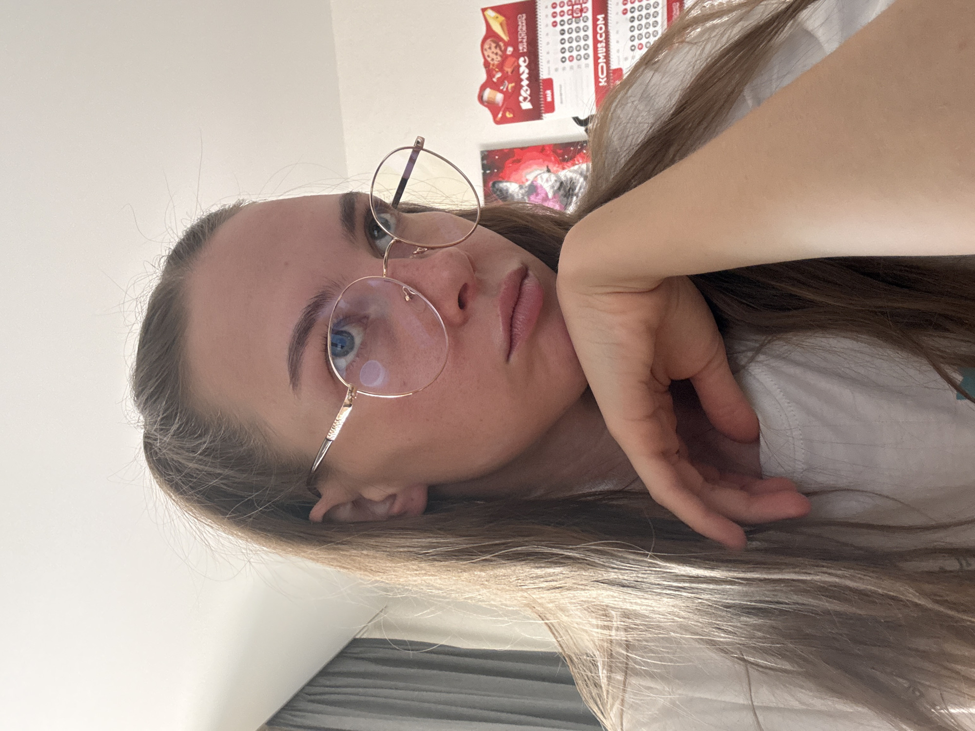 A young woman with long, light brown hair, wearing glasses, is resting her chin on her hand. She is looking slightly to the left with a neutral expression. Behind her, a grey curtain is visible on the left, and a calendar and a poster are on the right. The calendar has Russian text and the website "KOMUS.COM". The overall lighting suggests an indoor setting. The image does not contain any information that can be used to determine its location in the city of Perm, Russia.