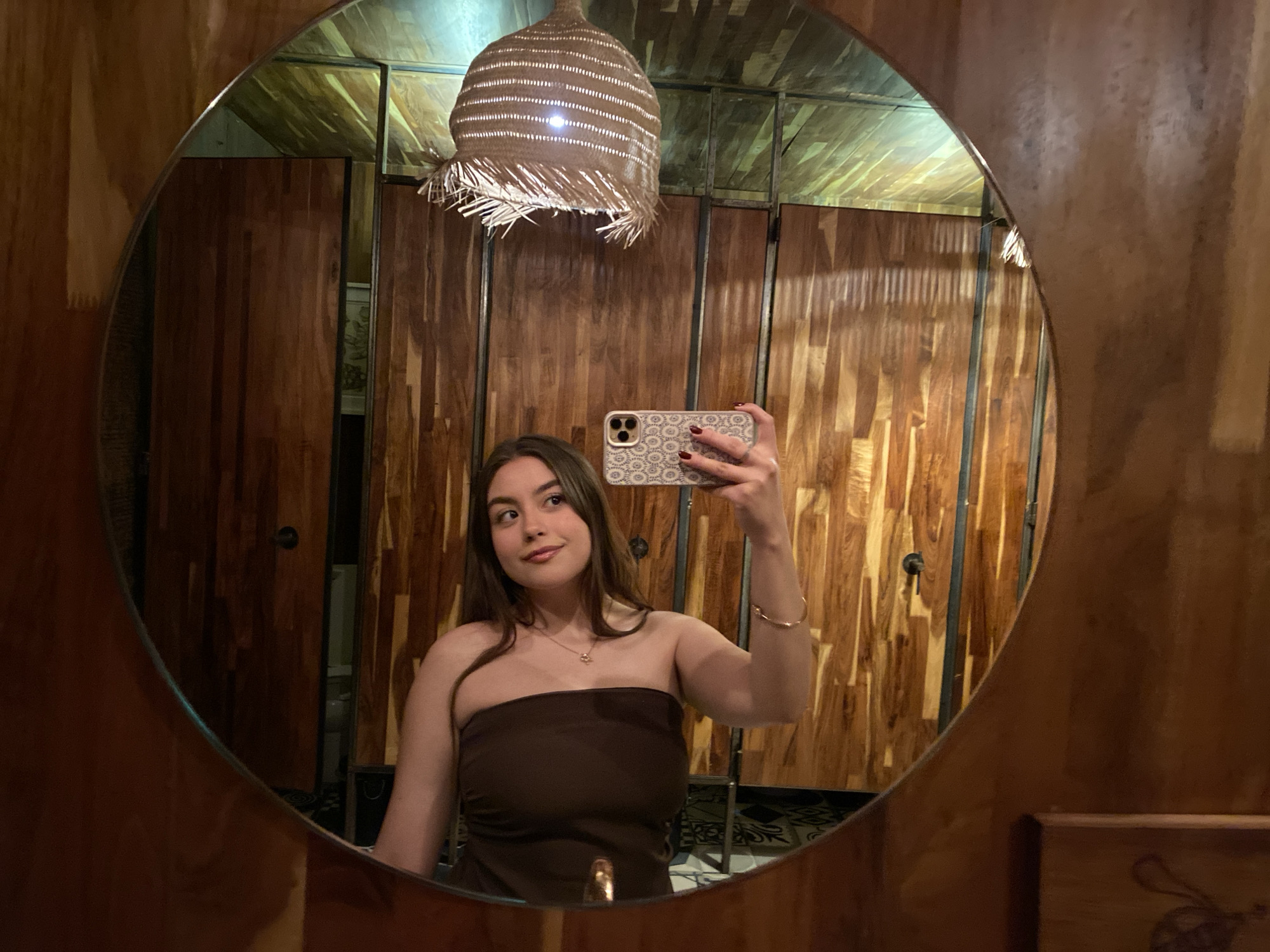 A medium shot depicts a young woman taking a selfie in a round mirror in a bathroom in Oaxaca City, Mexico. The woman has long brown hair, a brown tube top dress, and a gold bracelet on her right wrist. She is holding her iPhone in her right hand, its case decorated with a beige and white floral pattern. The bathroom features wood paneled walls and stalls, a woven light fixture hanging from the ceiling, and green accents on the ceiling. A glimpse of black and white patterned flooring can be seen in the reflection. A part of a sink is visible in the bottom.