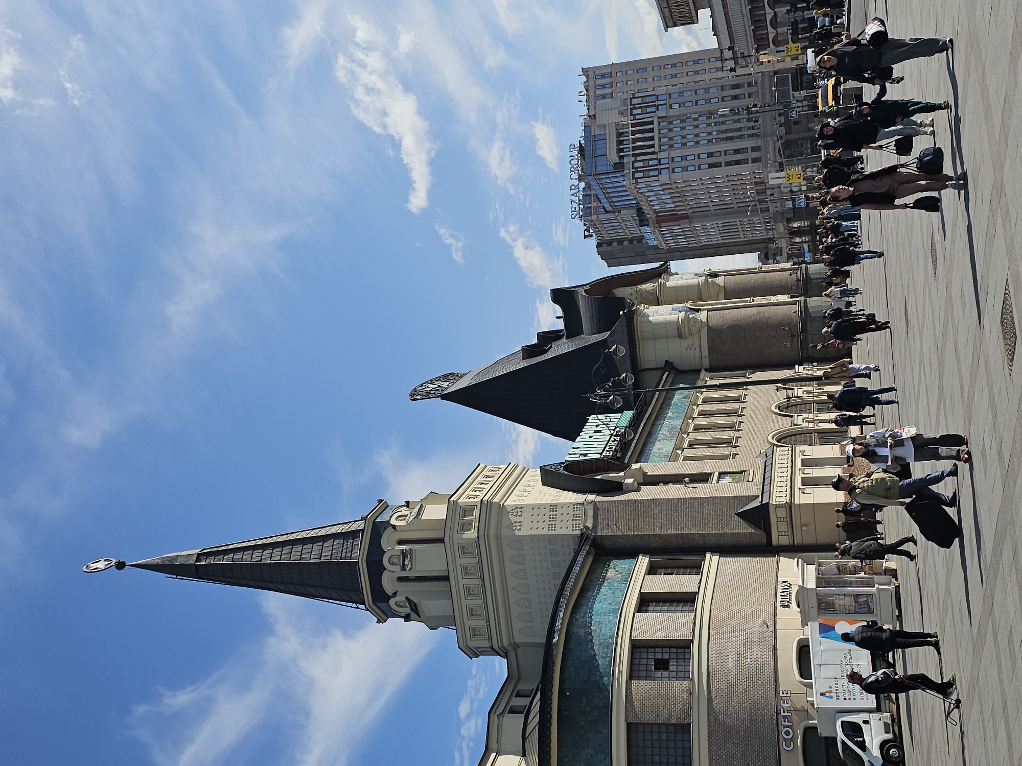 The image depicts a wide outdoor plaza in Moscow, Russia, in front of a grand architectural structure featuring a tall, dark spire topped with a circular emblem, likely a train station given the surrounding context of urban transit. The building exhibits intricate detailing with beige brickwork, ornate decorative elements, and turquoise tiled sections. In the background, modern high-rise buildings, including one with "SEZAR GROUP" signage, rise against a clear blue sky with wispy white clouds. A number of people are visible, walking across the paved plaza, some carrying luggage, suggesting travel or daily commute. A white van with Russian text and branding is parked to the left, and a small kiosk stands nearby. The overall scene captures a moment of urban activity with a blend of historical and modern architecture under a bright, sunny sky.