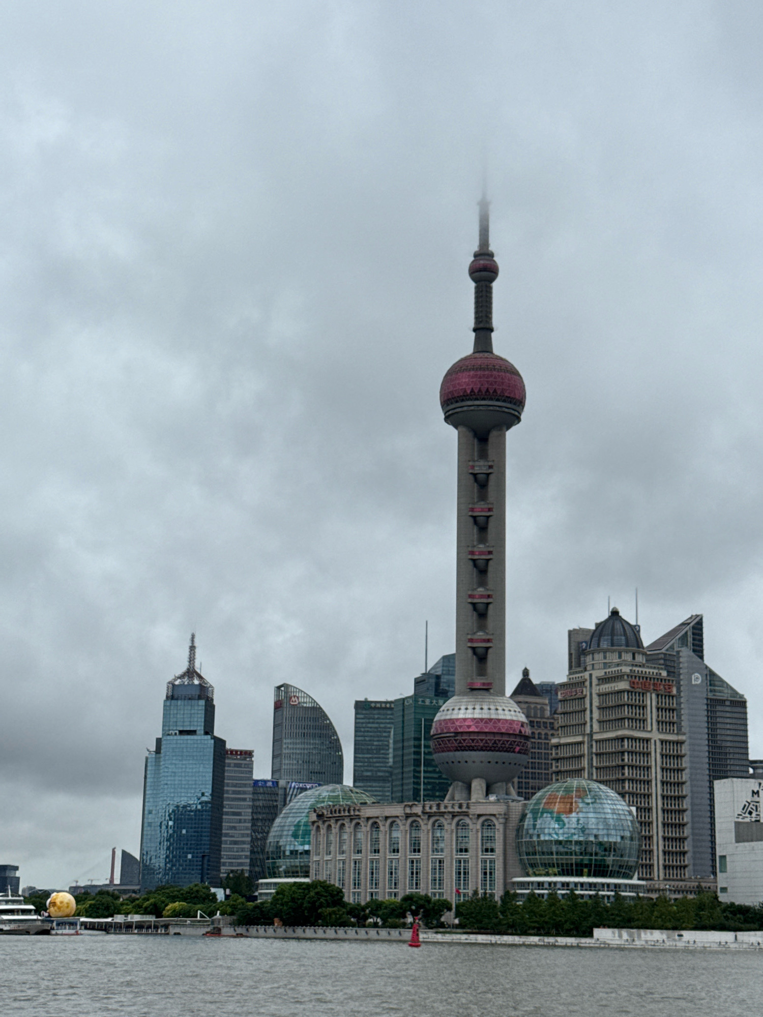 This image captures a cloudy day in Shanghai, China, focusing on the iconic skyline of the Lujiazui financial district. The Oriental Pearl Tower, a prominent landmark, stands tall with its distinctive spheres, partially obscured by mist at its apex. Surrounding it are various modern skyscrapers with distinct architectural styles, including the Shanghai World Financial Center and the Jin Mao Tower. In the foreground, a body of water, likely the Huangpu River, stretches across the frame. On the riverbank, a promenade with trees and a large spherical art installation resembling a globe are visible. A white boat or ship is docked on the left side of the river. The overall scene is characterized by a dense urban landscape under an overcast sky.