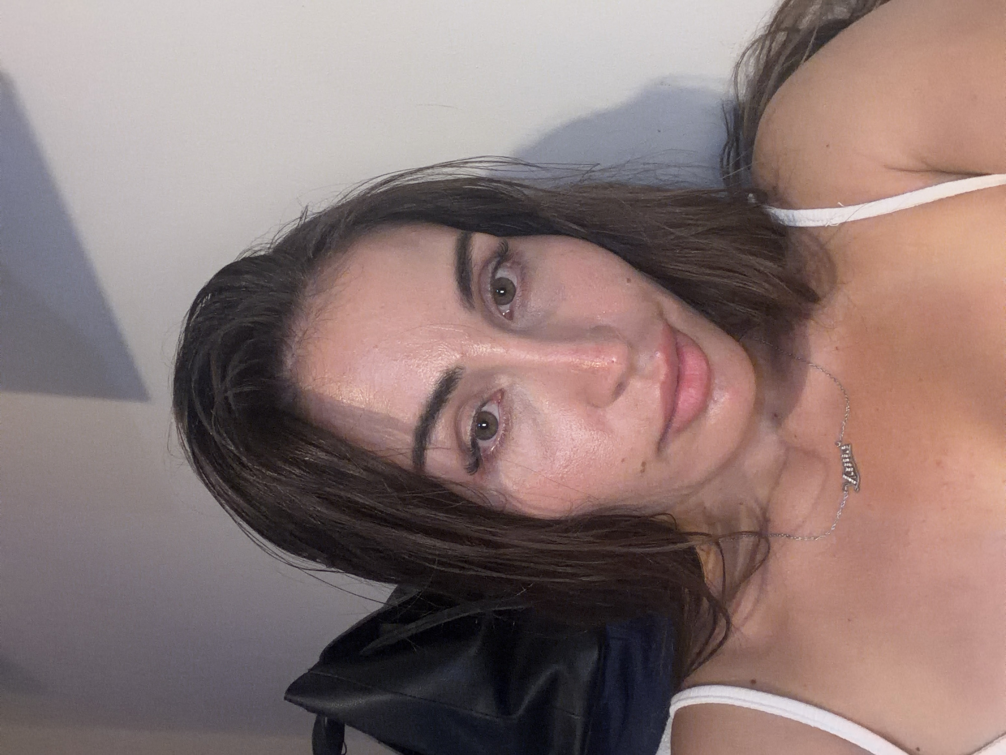 The image is a close-up selfie of a young woman with long, dark brown hair, taken indoors against a plain wall. She is wearing a white top and a silver necklace with a pendant that appears to have a name on it. Her skin is lightly tanned and appears to have some shine, possibly from sweat or makeup. The lighting suggests it might be evening or dimly lit. There is a dark bag on the left side of the frame, behind her. There is no visible indication of the city of Tersefanou, Cyprus, in the image.