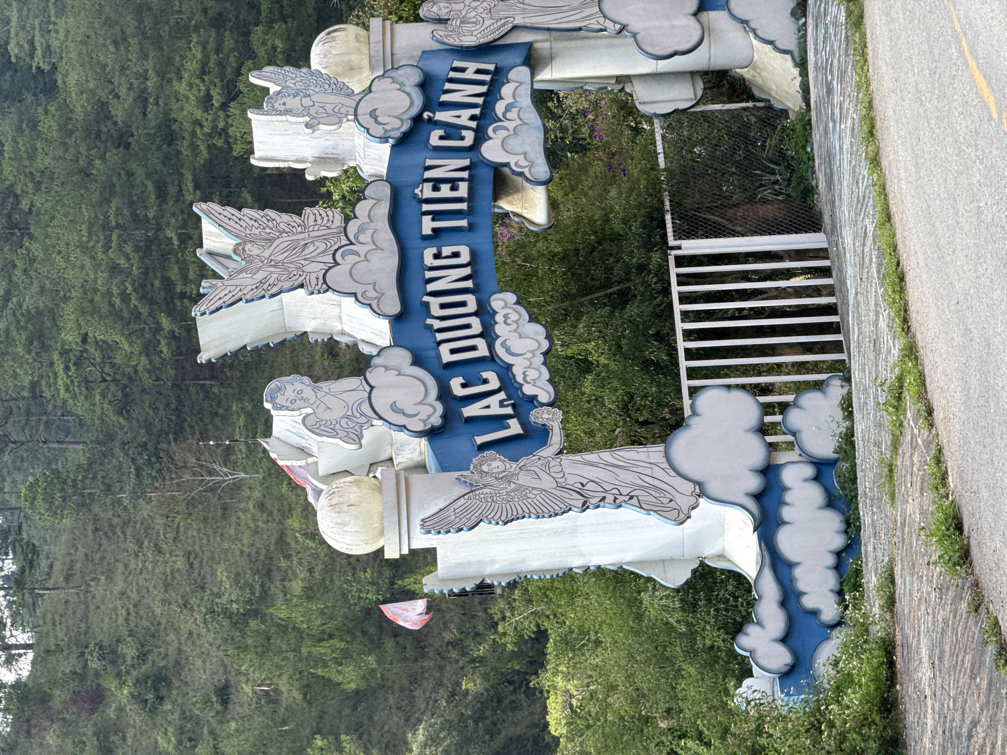 The image captures an entrance sign in Đa Nghịt, Vietnam. The sign, adorned with stylized angels and cloud motifs, reads "Lạc Dương Tiên Cảnh" in vertical lettering. The sign is bordered by dense green foliage and positioned adjacent to a paved road. A metal fence is visible in the background. The overall setting suggests a natural or scenic area, likely a park or tourist destination.