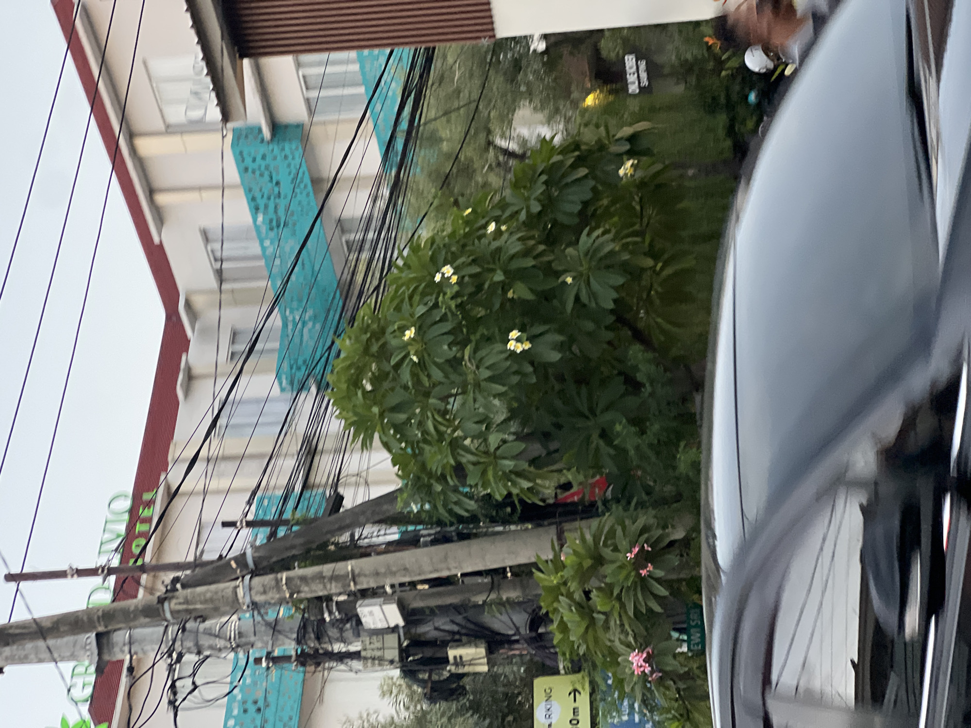 In Kuta, Indonesia, a street view captures a hotel with a red roof and white walls, adorned with turquoise decorative panels. A tall utility pole, heavily laden with electrical wires, stands in the foreground, partially obscuring a lush green tree with white and pink flowers. To the left of the utility pole, a yellow sign reads "PARKING" with an arrow pointing to the right and "50m". A dark-colored car moves through the foreground, its reflection visible on its glossy surface. Further in the background, a wooden-paneled building is visible to the right. A street sign with green text reads "EWIK SR".