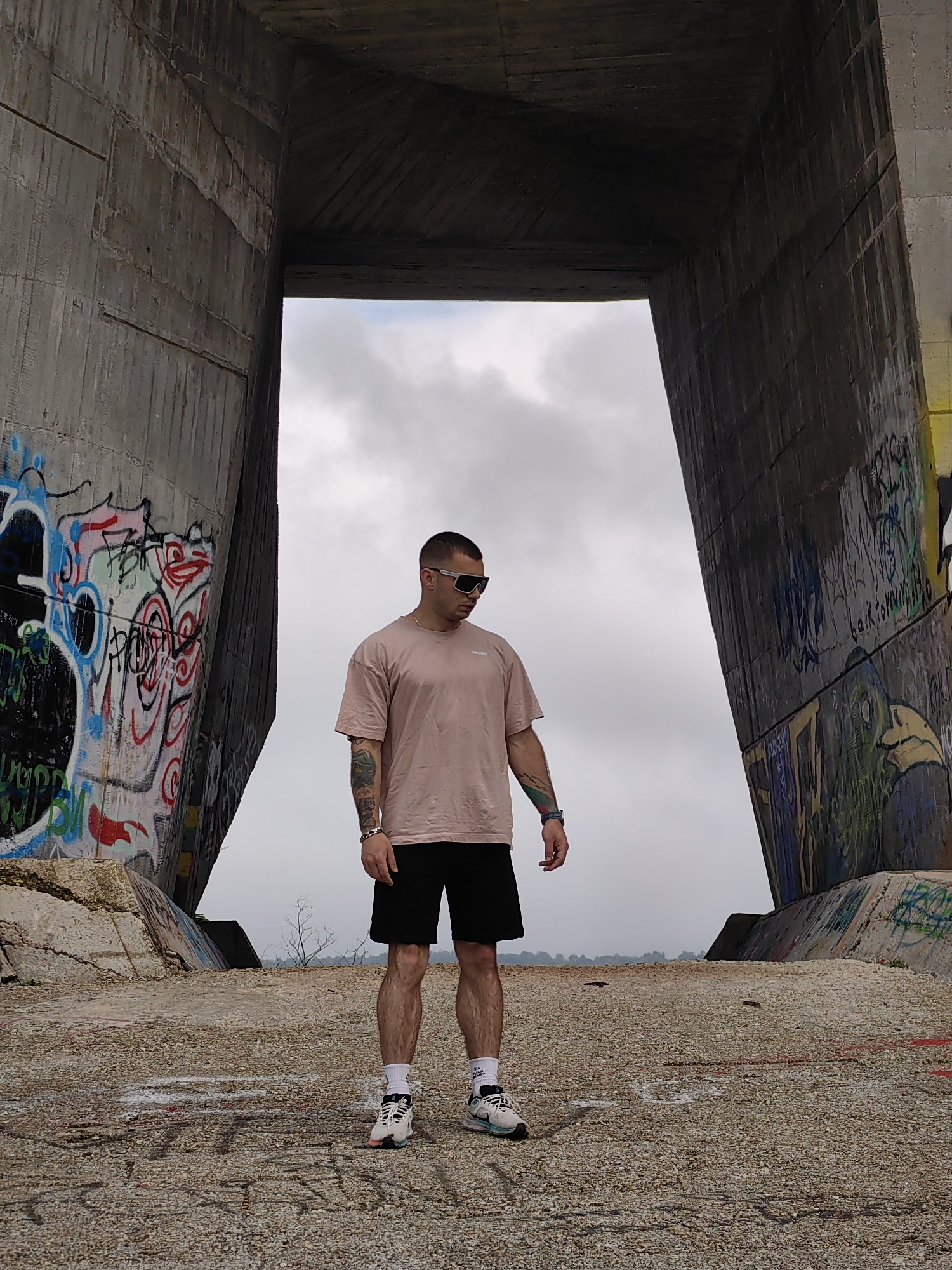 The image depicts a man standing under a concrete bridge structure in Varna, Bulgaria. The man is wearing a pink t-shirt, black shorts, white socks, and athletic shoes. He has short hair, sunglasses, and tattoos on his arms. The concrete structure features graffiti on its walls and a cloudy sky is visible through the opening. The ground is covered in loose gravel and more graffiti.