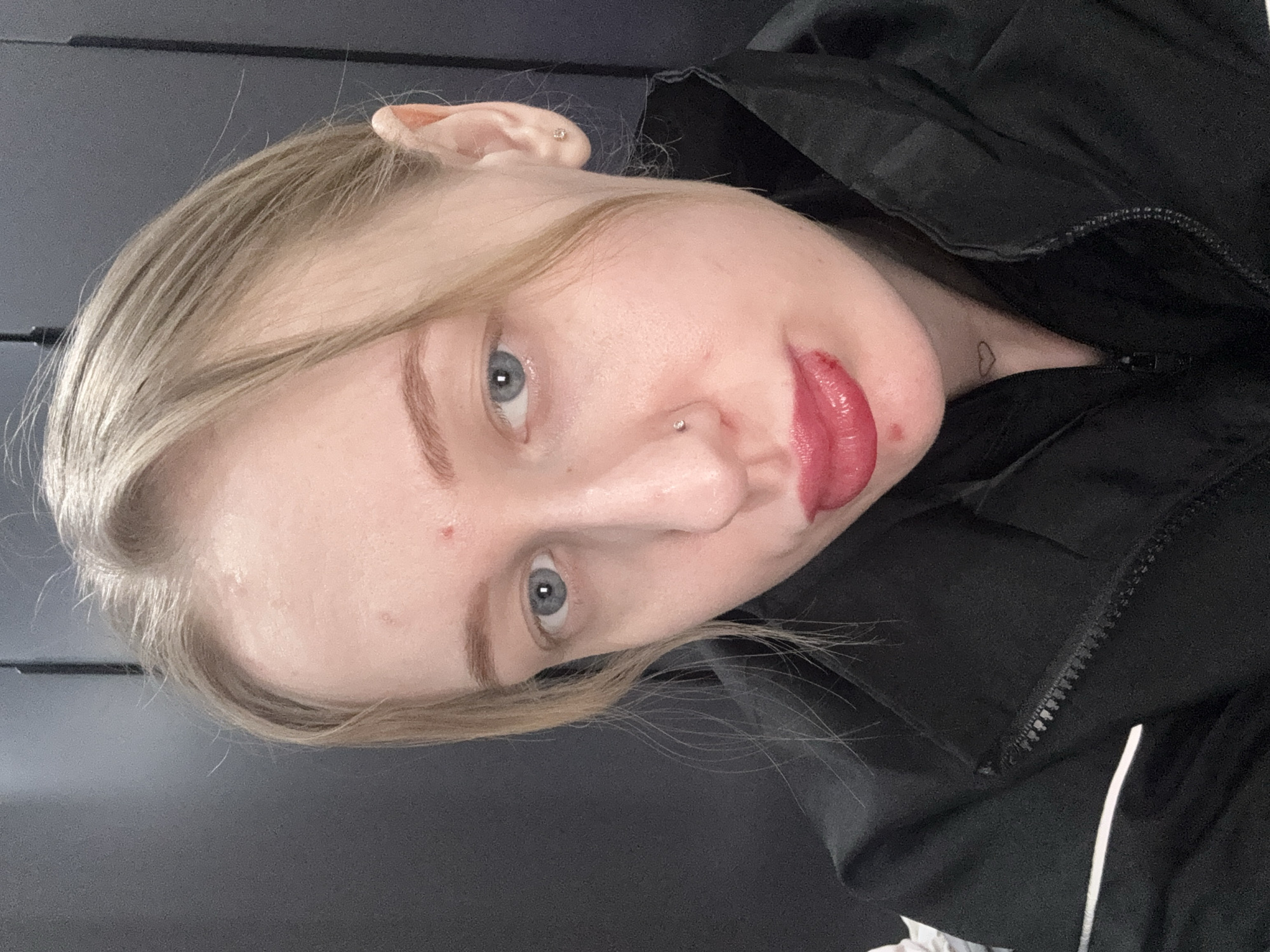 A young, light-skinned individual with blond hair and blue eyes is photographed indoors. They have a nose piercing and are wearing red lipstick. A small heart tattoo is visible on their chest. The individual is wearing a black jacket with a white stripe. The background is dark gray. There is no information about the location in Novoaleksandrovka, Russia, so I cannot include that.
