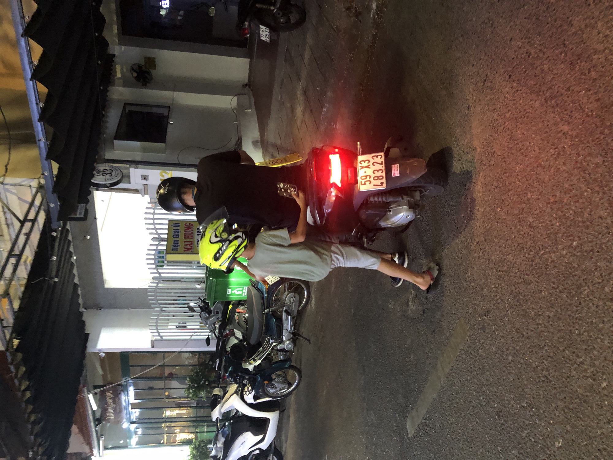 This is a nighttime photograph taken on a street in Đà Nẵng, Vietnam, showing a scene with several motorcycles and people. The image is oriented vertically, with the top of the frame appearing on the left.
In the foreground, two individuals are visible. One person, wearing a black shirt and helmet, is seated on a dark-colored scooter. They are holding a smartphone with a patterned case. Next to them, a younger person, wearing a light gray t-shirt and shorts, is standing or leaning. They are wearing a bright yellow and black helmet and have their right leg extended. Their left leg is bent at the knee, and they are wearing sneakers.
Several motorcycles are parked or present in the scene. The scooter in the foreground has a license plate that reads "59-X3 483.23". Another motorcycle behind it has a license plate that appears to say "77-61 23531". Further back, there are more motorcycles and a white scooter.
The setting appears to be an urban environment with buildings lining the street. A dark, corrugated awning extends over the sidewalk on the left. Signs are visible on some of the buildings, including one with Vietnamese text that reads "Tiệm Giặt Ủi MAI HƯNG". Another sign says "Little Georgia". The ground is paved with asphalt. The lighting suggests it is evening or night, with artificial lights from the buildings and the illuminated tail lights of the scooter. The overall atmosphere is casual, perhaps of people gathered on the street.