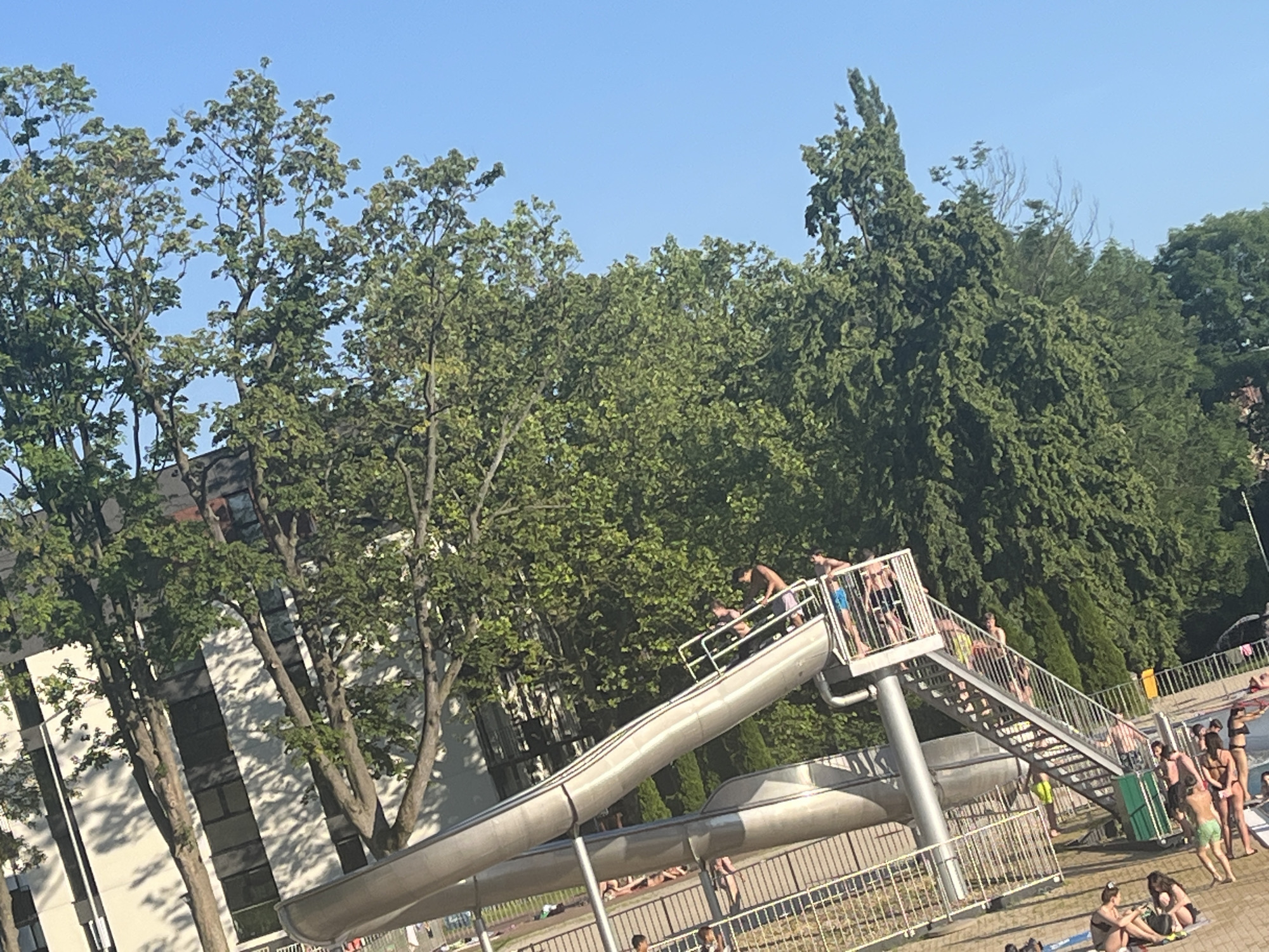 In the city of Unknown, Unknown, a sunny day is captured at an outdoor water park, with a clear blue sky overhead. The foreground features a large, winding water slide, its metallic structure curving through the scene. A metal staircase, also part of the slide's infrastructure, ascends to the top. Several individuals are visible on and around the slide. Some are at the top of the stairs, preparing to descend, while others are on the slide itself. A group of people are gathered near the bottom of the slide, some standing, others sitting on the ground. In the background, lush green trees form a dense canopy, with a building partially visible behind them on the left. A chain-link fence runs along the bottom of the image, separating the water park area from a paved walkway. The overall impression is one of a recreational activity on a warm day.
