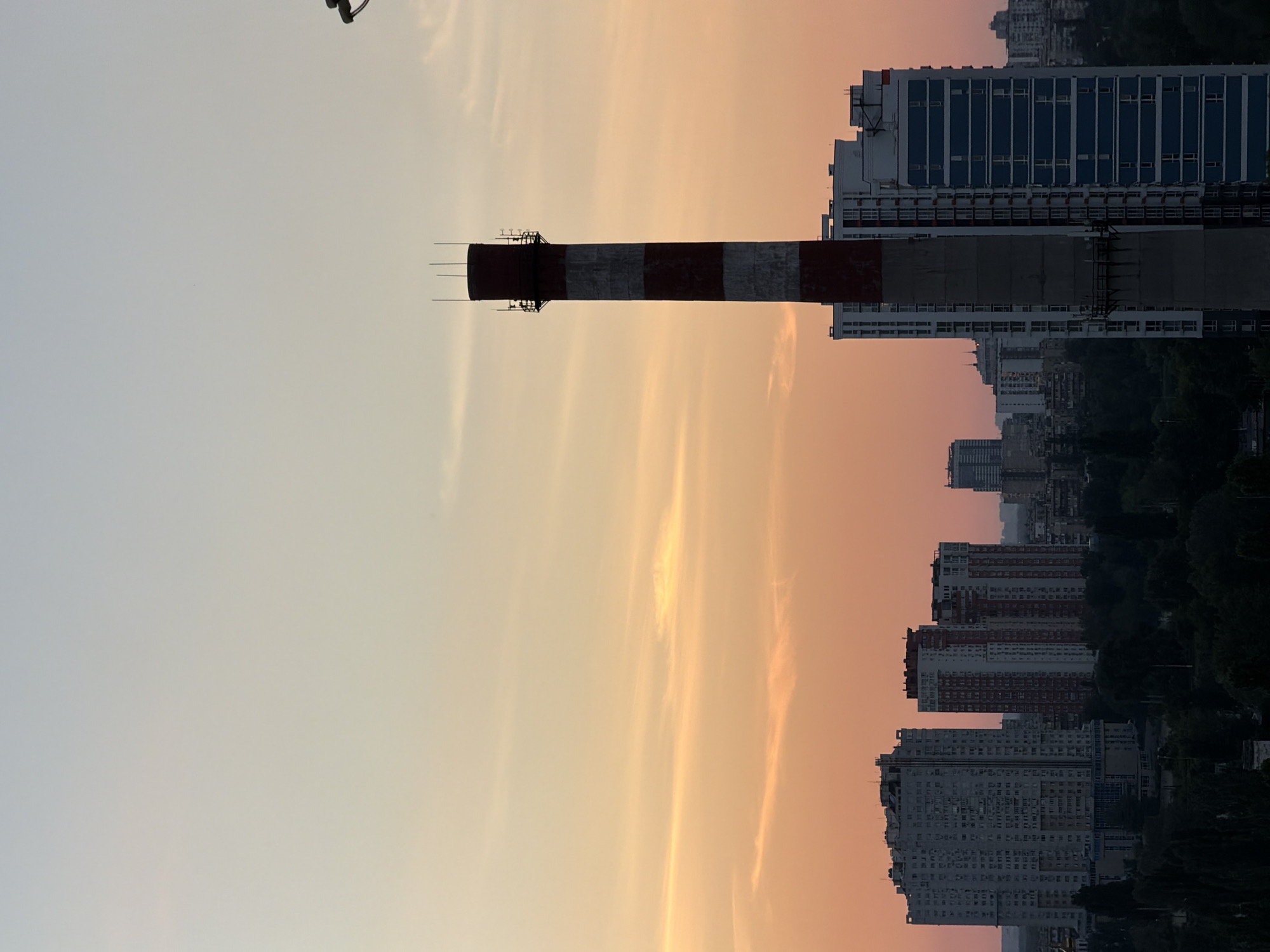 The image depicts an industrial chimney with red and white stripes against a soft, pastel-colored sky. The sky transitions from a pale blue at the top to a warm orange and pink hue, indicating either sunrise or sunset. Wispy clouds are scattered across the sky, adding to the atmospheric effect.
To the right of the chimney, a cityscape of tall residential buildings is visible. These buildings are modern, with many windows and balconies. Some of them are adorned with blue and white facades, while others are lighter in color. In the background, more buildings can be seen, receding into the distance and partially obscured by trees, suggesting a densely populated urban environment. The overall scene conveys a sense of urban living juxtaposed with industrial presence.
The orientation of the image is rotated, with the chimney appearing to stand vertically from the bottom of the frame. The lighting suggests it is either early morning or late evening. No people are clearly visible in the image. There is no visible text in the image. The location is identified as Kyiv, Ukraine.