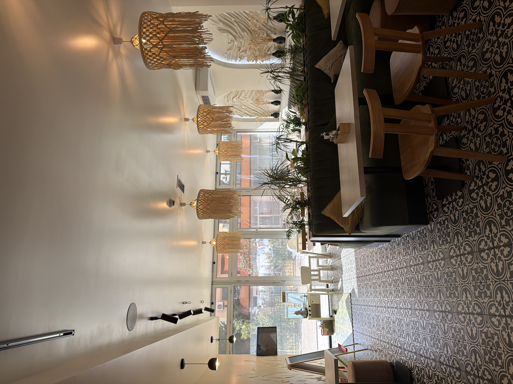 The image depicts the interior of a cafe or restaurant, oriented vertically. The scene showcases a modern and cozy ambiance with a focus on natural materials and decorative lighting. Several macrame-style pendant lamps with fringe accents hang from the ceiling, casting a warm glow. The walls are a light, neutral color, providing a backdrop for the various elements.
In the foreground, a long, dark green sofa runs along the wall, adorned with throw pillows in light beige and cream. In front of the sofa, white rectangular tables are visible. Wooden chairs with curved seats and backs are positioned around the tables, some of which are partially obscured.
A prominent feature is the patterned tiled floor, a dark gray with an intricate white design, adding a graphic element to the space. Large windows and glass doors allow natural light to enter, revealing an outdoor area with greenery and glimpses of buildings.
Other objects in the scene include a television screen mounted on a stand, and various lamps and light fixtures along the wall. A few potted plants are strategically placed, contributing to the relaxed atmosphere.
The overall impression is a well-lit, contemporary indoor dining or seating area, possibly during the daytime given the natural light. There are no visible people actively engaged in activities, though the setting suggests it is prepared for customers. The style is consistent with a trendy eatery or lounge.