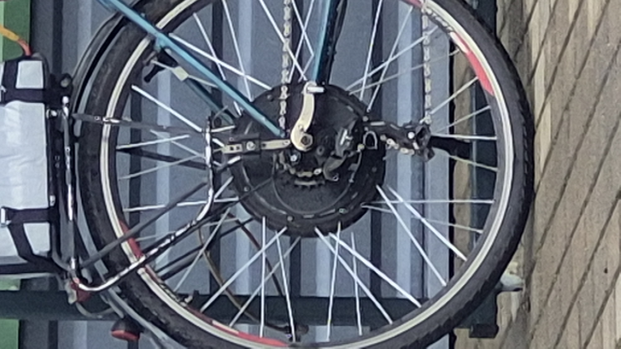 A close-up, slightly blurry, eye-level shot shows the rear wheel and drivetrain of a bicycle parked outdoors. The bicycle has a black, circular hub with a metallic silver derailleur attached. A silver chain extends from the derailleur towards the right of the frame. The wheel itself has numerous thin, silver spokes and a black tire with a red stripe. A black metal rack is attached above the rear wheel, supporting a light grey bag. The background consists of a muted blue, horizontal ribbed surface, possibly a wall or fence. The ground in the foreground appears to be a light brown, paved surface with distinct parallel lines, likely wooden planks or bricks. The scene is static, with no people or animals visible. This bicycle is located in the city of Pabradė, Lithuania.