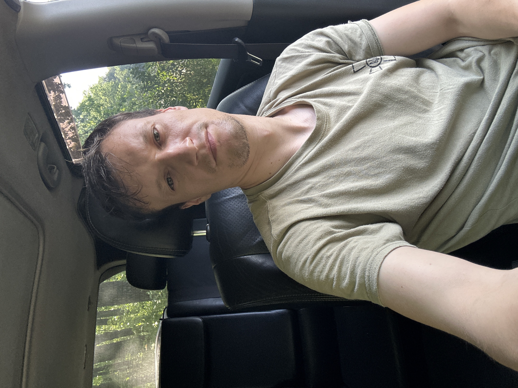 A man is shown from the chest up, sitting in the passenger seat of a car. He is looking directly at the camera. The interior of the car is dark, with black leather seats visible. Outside the car windows, trees and foliage can be seen, suggesting a rural or wooded location. The man is wearing a light olive-colored t-shirt with a black emblem on the right sleeve. The image is well-lit, with natural light coming in from the windows. There is no indication of the specific location within Makiv, Ukraine.