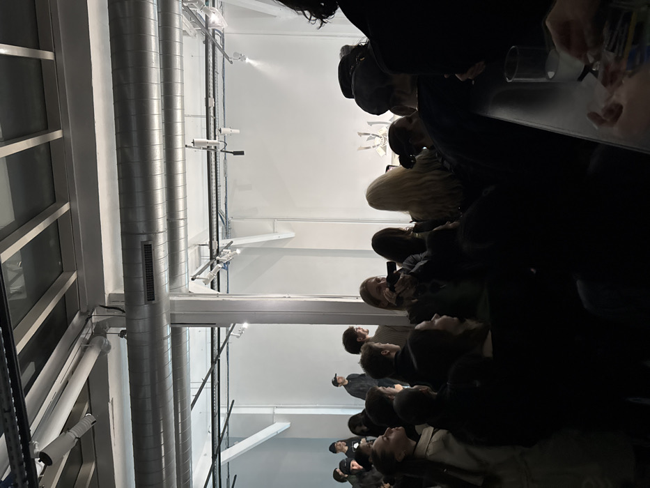 The scene depicts an indoor event within a modern industrial-style exhibition space in Paris, France. A standing crowd of approximately twenty to thirty individuals, predominantly young adults, occupies the foreground and mid-ground, facing towards a brightly illuminated central display area. The venue features white walls, exposed silver cylindrical air ducts, and overhead black track lighting fixtures with spotlights. In the background, a white, multi-faceted, abstract sculptural object or display is prominently positioned under direct lighting. A person with blonde hair in the mid-ground, slightly left of center, is holding a black camera and photographing the display. On the far right foreground, a clear glass containing liquid rests on a dark surface, and several hands are visible interacting with small, indistinct objects. The foreground is dimly lit, contrasting with the intense illumination of the background display. Two males wearing caps are observable standing in the far left background.