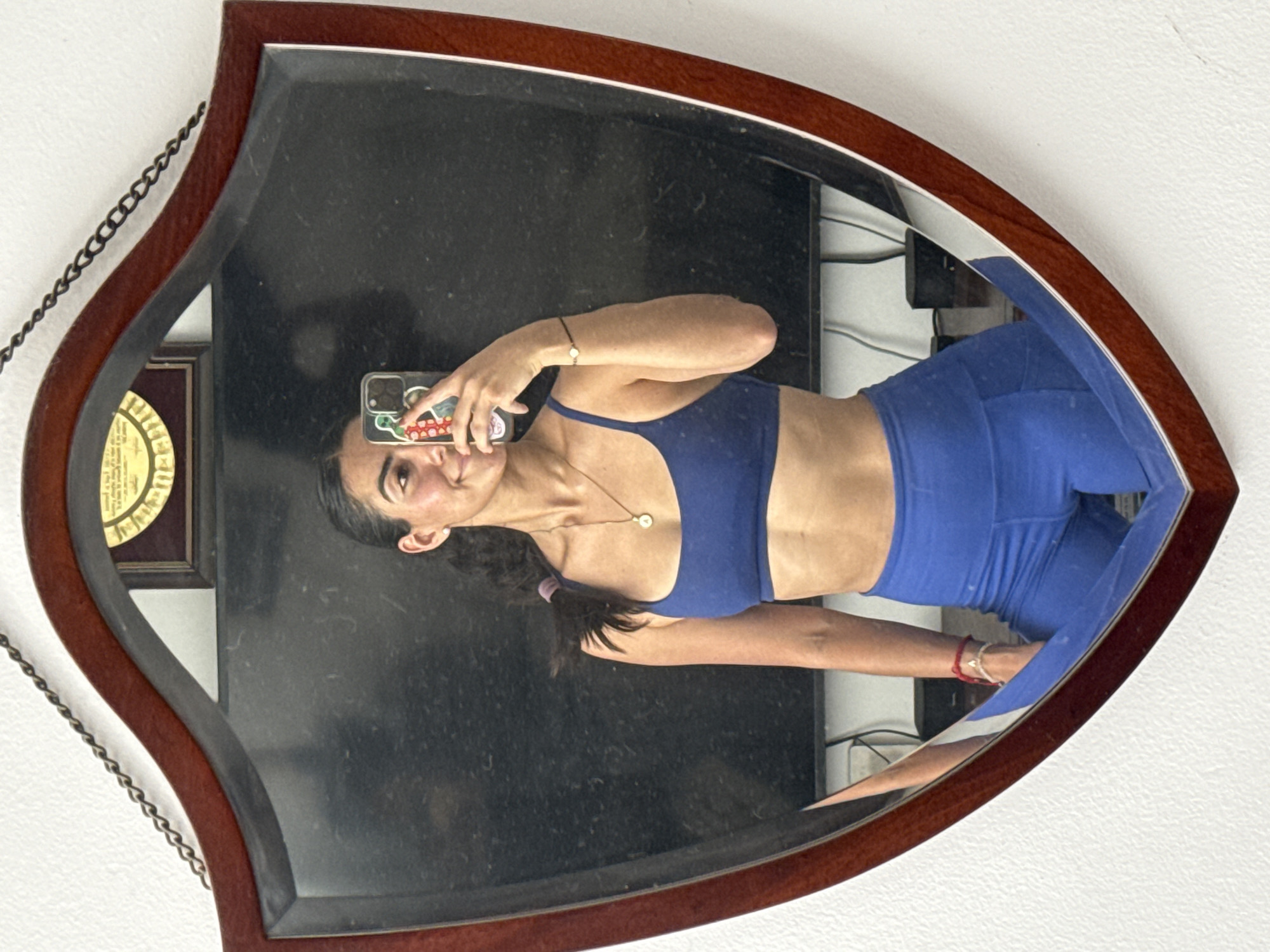 A young woman in a blue sports bra and blue leggings takes a selfie in an ornate, teardrop-shaped wooden-framed mirror. A certificate hangs on the wall to the left of the mirror. A dark television is visible in the reflection. A white wall is visible around the edges of the mirror. The location is likely indoors, possibly a home or apartment, but the city of Puebla, Mexico cannot be confirmed.