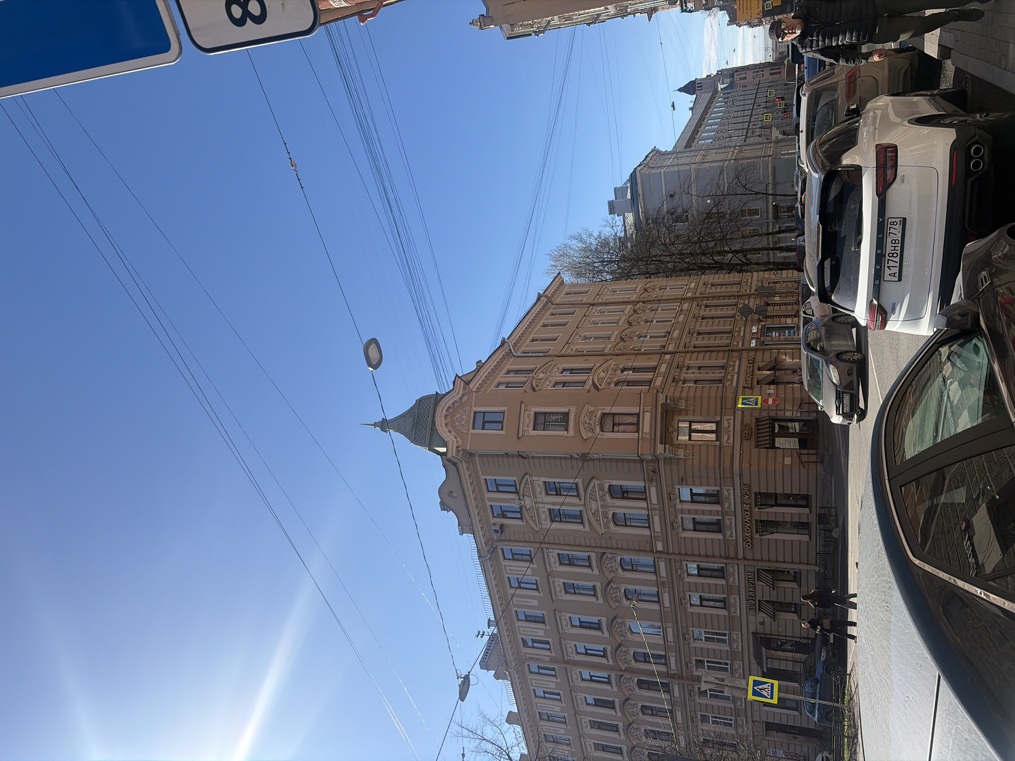 This media file captures a sunny daytime street scene in Saint Petersburg, Russia. The perspective looks along a city street lined with buildings and parked cars.
The most prominent subject is a multi-story building on the left, featuring a light brown/beige facade with ornate architectural detailing around its numerous windows and cornices. A distinctive green-roofed, pointed turret or dome crowns one of its corners. Bare tree branches are visible in front of this building, suggesting late winter or early spring. Several signs with Russian text are visible on the ground floor, including "Будочная №6" (Booths No. 6) and "Кофенхаузъ" (Kofenhaus/Coffee House).
On the right side of the street, a line of cars is parked. A white Geely SUV is clearly visible, bearing the Russian license plate "A178HB 778," which indicates the Saint Petersburg region (778). A person in a dark jacket and sunglasses stands near this car. Another dark vehicle is partially visible in the immediate foreground. Further down the street, two figures are walking on the sidewalk near a yellow pedestrian crossing sign.
The weather is bright and clear, with a vivid blue sky. Overhead utility wires crisscross the sky, and a bright sun flare is visible in the lower left portion of the frame. Streetlights are also part of the urban infrastructure. The scene portrays an everyday street atmosphere in a European city setting.