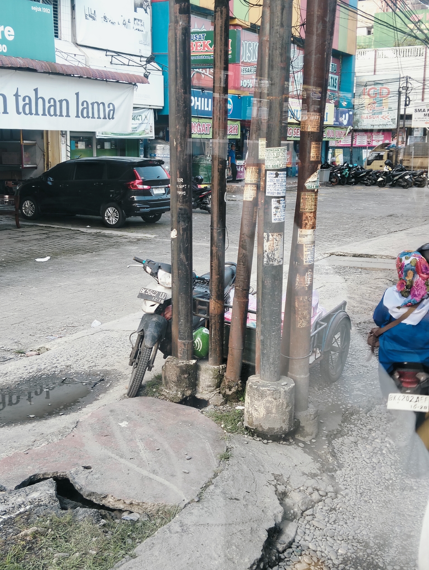 The image captures a street scene in the City of Medan, Indonesia, characterized by a mixture of commercial signage, vehicles, and infrastructure. In the foreground, a damaged concrete slab with exposed rebar and grass growing around it indicates a state of disrepair on the roadside. Several utility poles, weathered and covered in peeling posters, dominate the center of the frame. A motorcycle with a sidecar and a license plate reading "BK 5642 UU" is parked beside one of these poles. Behind the motorcycle, a black SUV is parked. Further in the background, a row of motorcycles is visible. Numerous colorful signs with Indonesian text, advertising various businesses and services, line the buildings along the street. A person wearing a blue jacket and a colorful headscarf is seated on a motorcycle, facing away from the camera. The overall environment appears to be an urban commercial area with ongoing street activity.