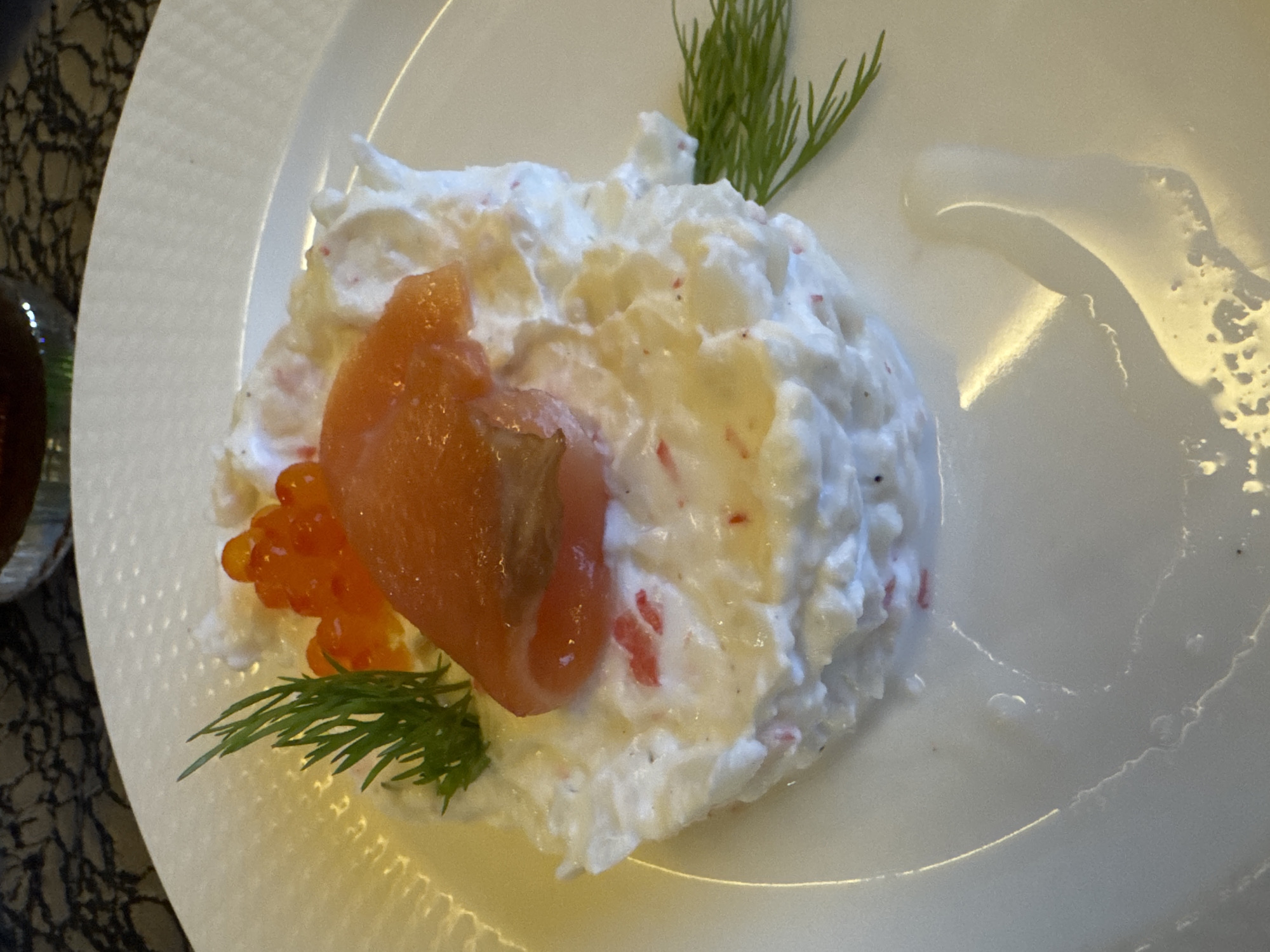 The image shows a plate of food. The main component is a white, fluffy mound, possibly a type of salad or mousse, speckled with reddish flecks. On top of this mound is a piece of salmon, some orange roe, and two sprigs of dill. A streak of liquid is visible on the right side of the plate. A partially visible glass is in the bottom left corner. The plate is white and appears to be part of a larger dish setting. The image location is unknown in Plovdiv, Bulgaria.