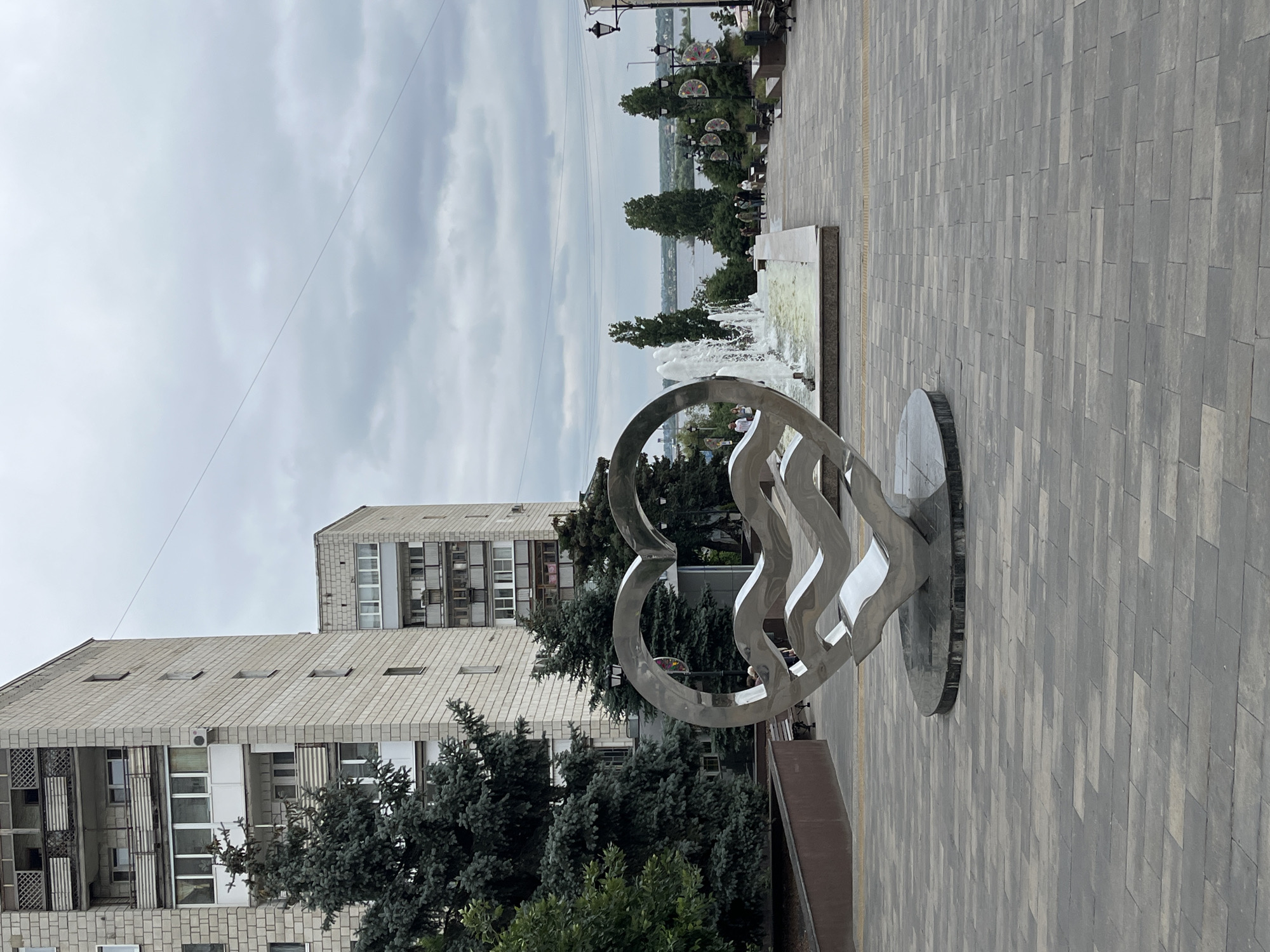 The image depicts a public plaza with a large, abstract stainless steel sculpture in the shape of a heart with wavy elements inside. The sculpture is situated on a paved walkway made of grey rectangular bricks. Behind the sculpture, a fountain sprays water upwards, and further back, a wide, paved pathway leads towards a body of water, likely the Volga River, with a distant cityscape visible under a cloudy sky. Several trees are planted along the pathway, and lampposts with decorative light fixtures line the sides. In the background, two tall, multi-story buildings constructed with light-colored brick are visible. A few individuals are scattered throughout the scene, walking on the pathway and near the fountain. The overall environment suggests an urban public space designed for recreation and aesthetics, possibly located in the city of Saratov, Russia, given the architectural style and the prominent river.