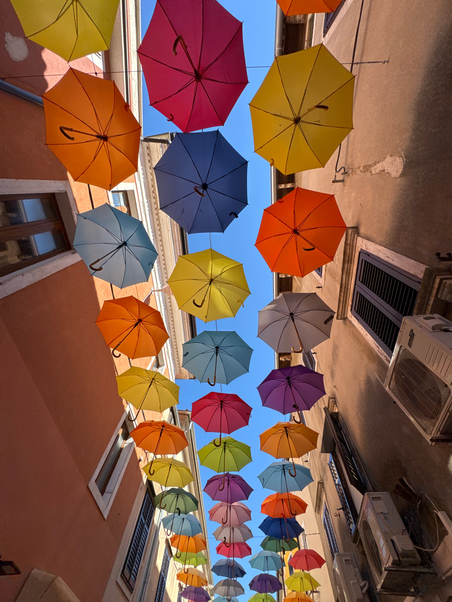 The image displays an overhead view of a street in Grad Novigrad, Croatia, adorned with a vibrant canopy of colorful umbrellas suspended across the narrow alleyway. The umbrellas, in shades of yellow, orange, pink, blue, and purple, are arranged in staggered rows, creating a visually striking ceiling against the clear blue sky. Buildings flank the street, their facades painted in warm, earthy tones, with visible windows and shutters. The perspective is from below, looking upwards, emphasizing the scale and impact of the umbrella installation. No people are visible in the frame, but the scene suggests a lively urban setting designed for aesthetic appeal.