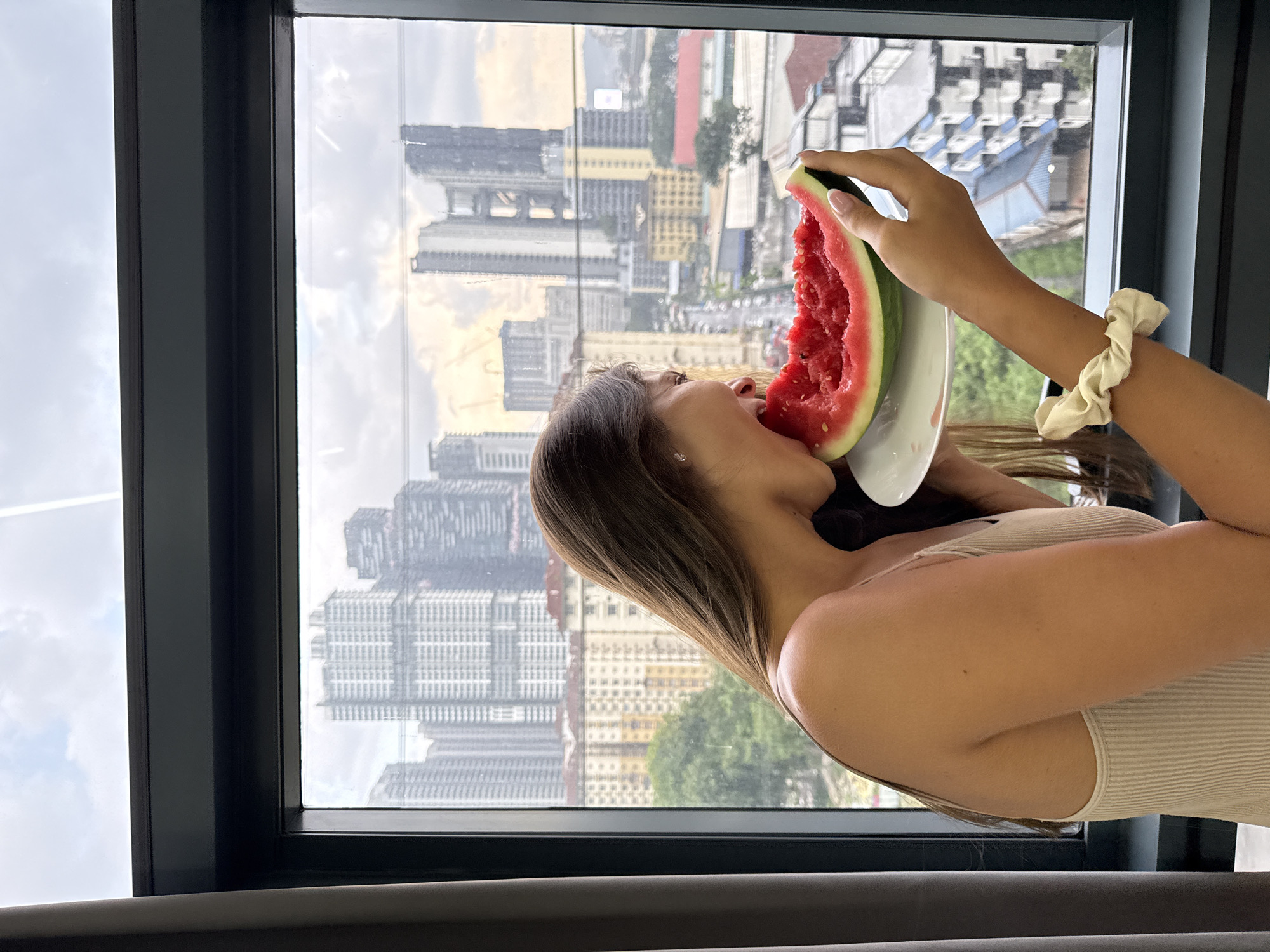 A medium-angle shot shows a woman in an indoor setting, leaning into a large window while eating a slice of watermelon. The image is tilted, giving the impression that the cityscape outside is rotated. The woman, with long brown hair, is seen from the mid-chest up, wearing a light-colored, ribbed tank top. She holds a thick slice of red watermelon, with visible seeds, to her mouth, supported by a white plate from underneath. A cream-colored scrunchie is worn on her right wrist.
Beyond the dark-framed window, a sprawling urban landscape of Kuala Lumpur, Malaysia, is visible under a bright, cloudy sky. Numerous high-rise buildings dominate the background, featuring diverse architectural styles, including several with grid-like window patterns. Some distant greenery and smaller buildings are also present. The scene is brightly lit, suggesting it is daytime. No discernible text is visible in the image. The woman's activity and the panoramic city view through the window create a relaxed urban scene.
