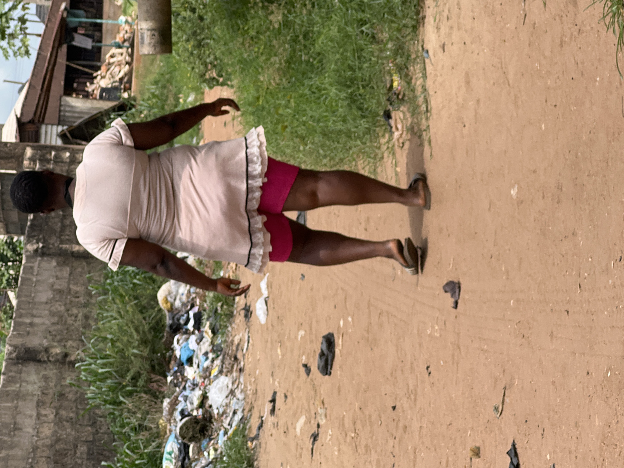 A woman is walking along a dirt road in Obosi, Nigeria. She is wearing a light pink dress with a white ruffle trim, and pink shorts underneath. She has dark skin and is wearing sandals. To her left, there is a pile of trash, grass, and a concrete wall. To her right, there is grass and a dirt road. In the background, there is a building with a corrugated metal roof.