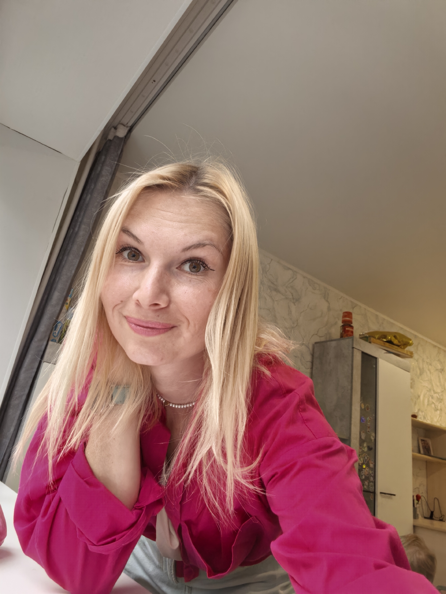A young woman with blonde hair and light brown eyes is pictured in a close-up, frontal shot. She is wearing a vibrant pink collared shirt and a pearl necklace. Her right hand is resting on her chin, and she is looking directly at the camera with a slight smile. The background appears to be an indoor setting, possibly a home, with a plain wall and a cabinet visible on the right side. There is no indication of the city of Sterlitamak, Russia, in the image.
