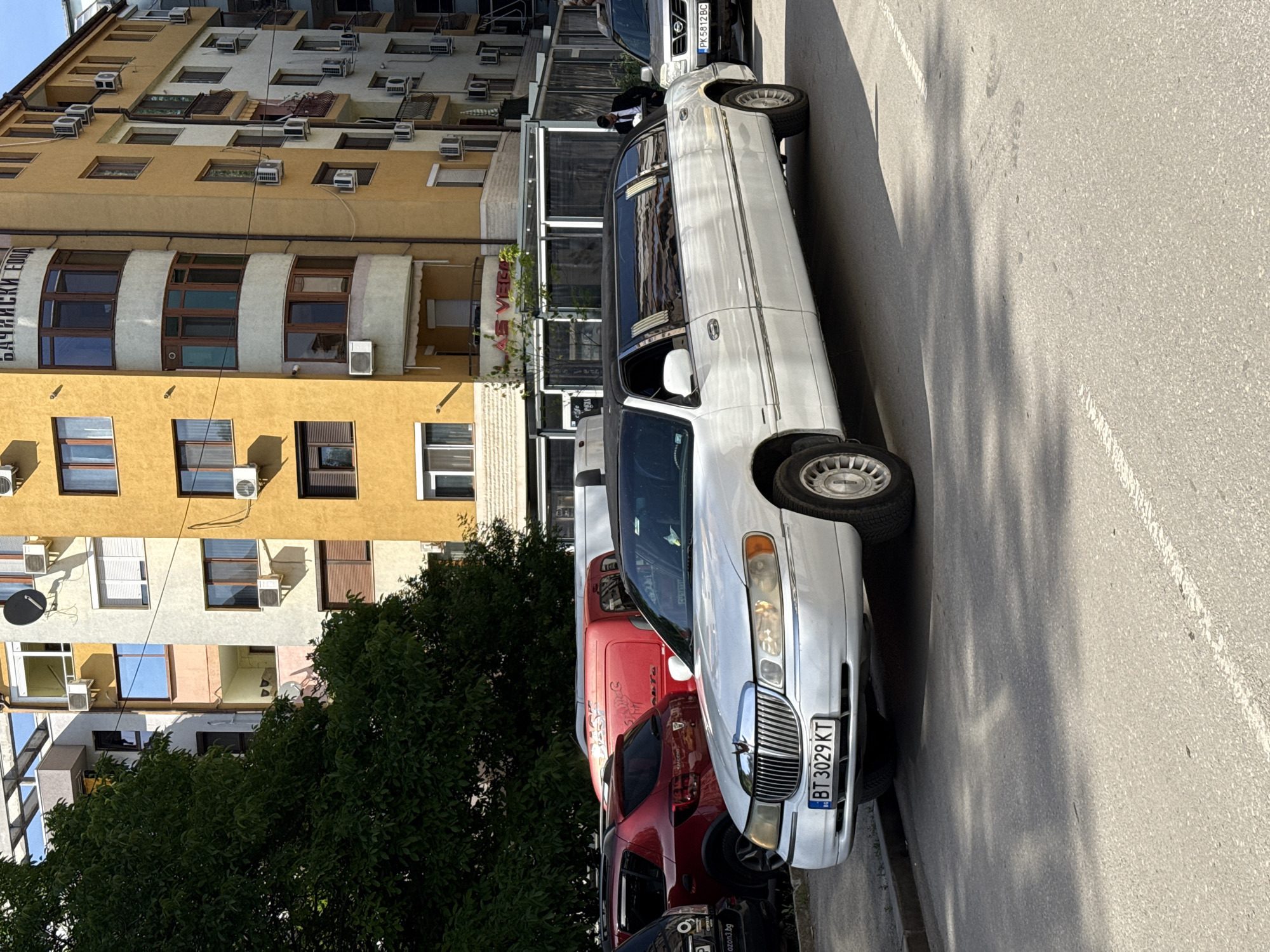 The image captures a street scene in Pleven, Bulgaria, featuring a white Lincoln Town Car limousine with the Bulgarian license plate BT 3029 KT parked along the curb. The limo is partially obscured by other parked vehicles, including a red van and a dark-colored SUV. The background consists of a multi-story building with beige and tan facades, characterized by numerous windows and air conditioning units. A dense green tree is visible to the left of the vehicles, partially obscuring the lower floors of the building. The street is paved with asphalt, and shadows from the trees and buildings are cast across the scene, indicating bright sunlight. The angle of the photo suggests it was taken from a slightly elevated position or a standing eye level. A sign for “AS VEGA” is visible above the parked van. The buildings in the shot appear to be apartments or hotels with satellite dishes.