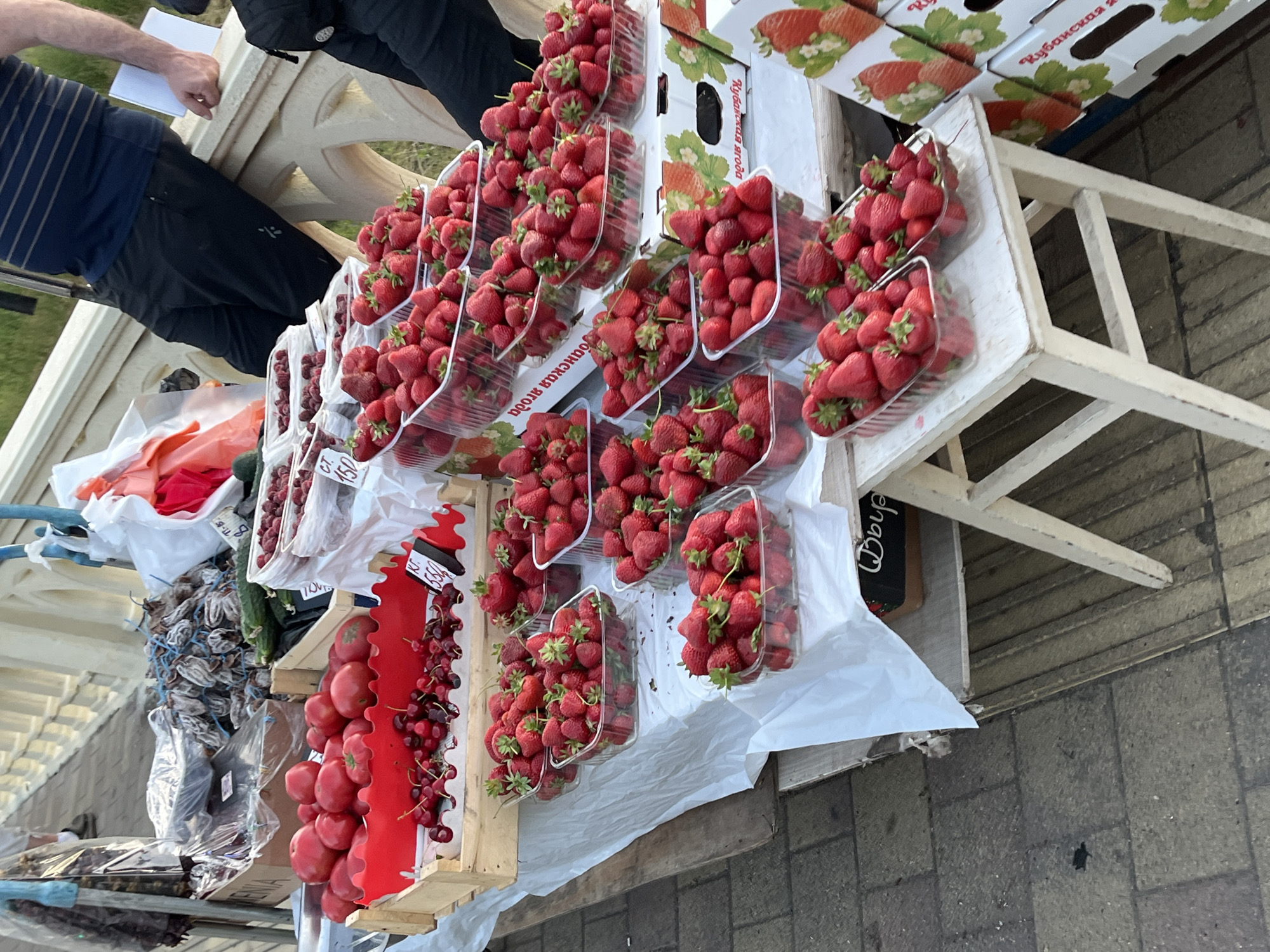 This is a medium shot, eye-level, exterior, daytime photograph of a fruit stand on a paved surface, likely a pedestrian walkway, possibly near the sea in Sochi, Russia. The fruit stand is laden with various fruits, predominantly strawberries and tomatoes, arranged in wooden crates and plastic containers.
In the foreground, several clear plastic containers filled with fresh, ripe strawberries are stacked and displayed on a white surface, some on a small, white wooden stool with visible wear and tear. To the left, a wooden crate holds a generous amount of bright red tomatoes, with a layer of dark red cherries above them. Behind the tomatoes, there are cucumbers and what appear to be dried fruits, possibly persimmons, piled in a plastic bag.
The background shows a partial view of a balustrade, suggesting the location might be a promenade or a bridge. Two individuals are partially visible. On the right, a person wearing dark pants and a striped t-shirt leans against the balustrade, holding a white piece of paper. Another person, partially visible on the far right, wears dark clothing and a backpack. A vendor's cart, with blue poles, is visible to the left of the frame, suggesting the operation of a mobile market.
The overall scene depicts a vibrant market stall offering fresh produce. The lighting suggests it's either early morning or late afternoon, with soft, natural light. The ground is paved with textured gray tiles. Signs with Cyrillic lettering are visible on some of the fruit packaging.