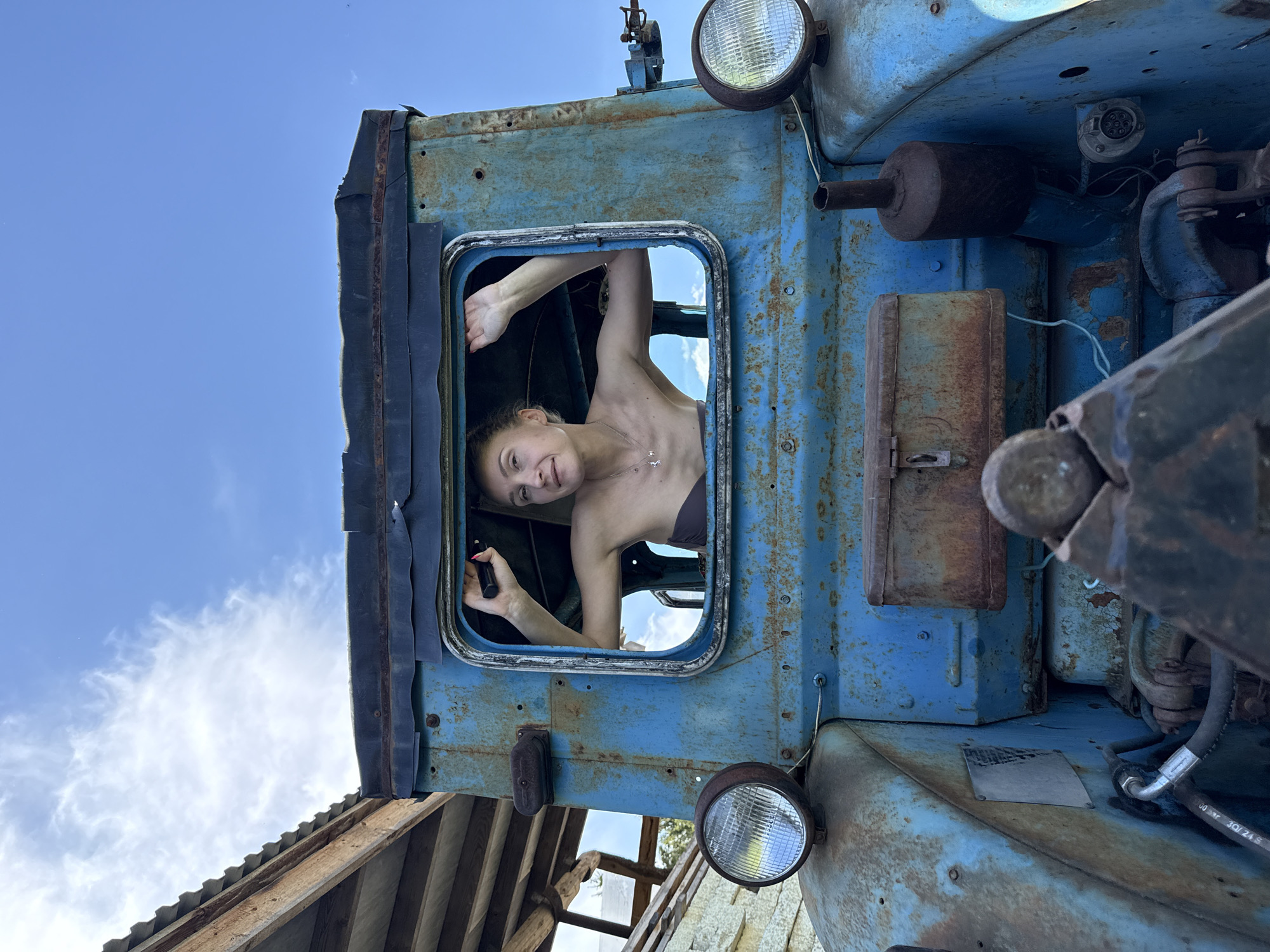 A person is visible through the window of a blue, old, and rusty vehicle. The person appears to be female with her arms extended upwards, holding a black object in one hand. The vehicle is positioned in an outdoor environment with a view of the sky, possibly in a rural or industrial location. Some parts of what appears to be a building's roof are visible in the lower left of the image. Based on the provided information, the location of the image is speculated to be Nizhnyaya Chermoda, Russia.