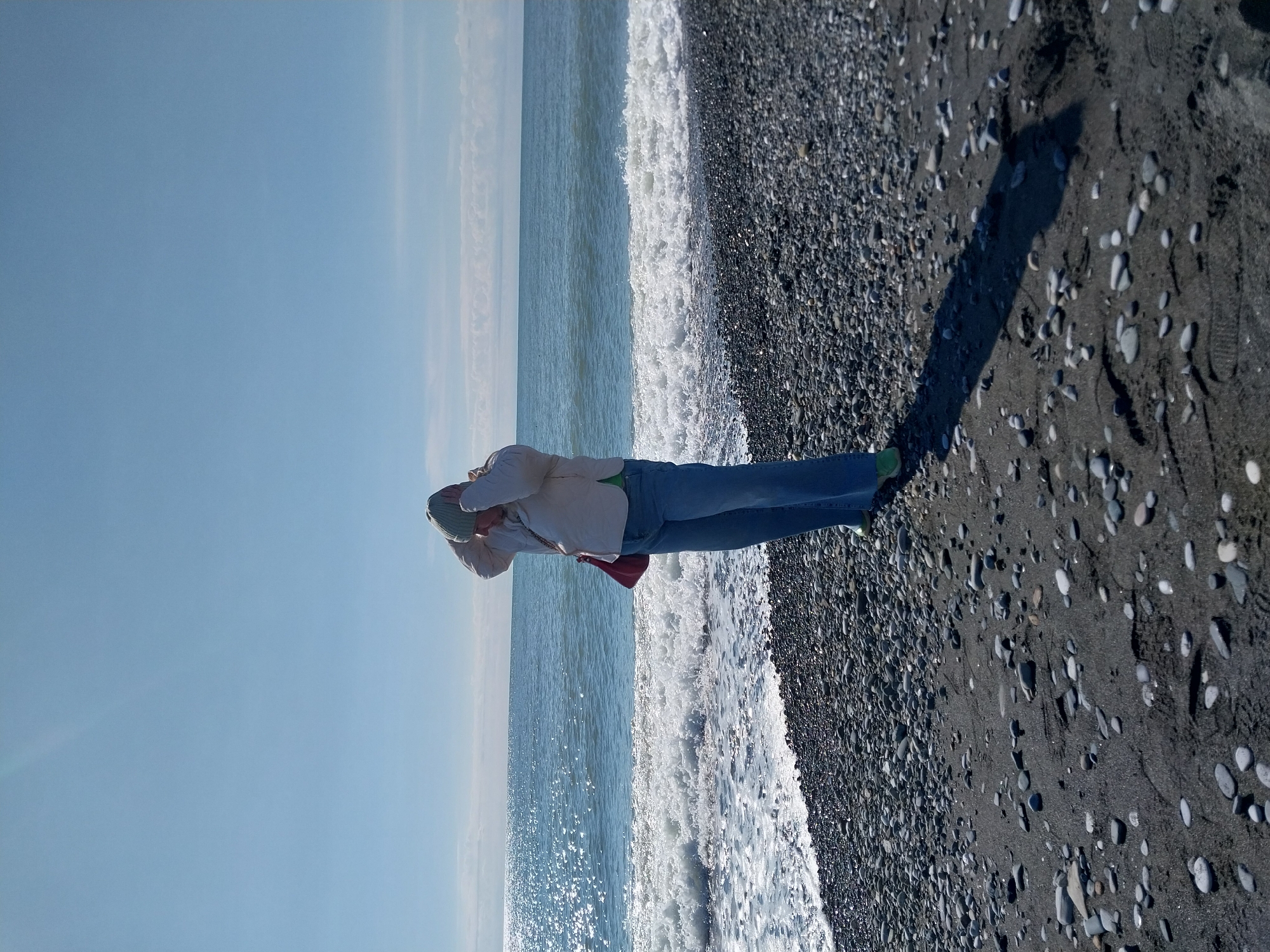 A woman stands on a pebble beach beside the sea in Sochi, Russia. She wears jeans, a light-colored jacket, a beanie, and possibly green shoes. She is holding her hands on her head. A red purse is visible. The waves are breaking on the shore, and a shadow stretches across the pebbles. The sky is clear and blue.
