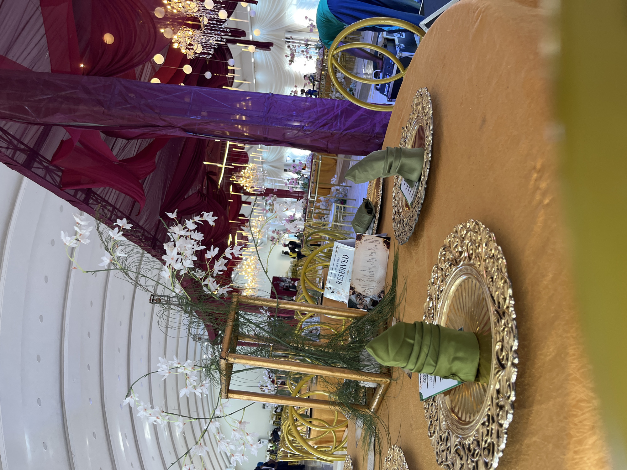 The image shows a decorated event space, possibly a banquet hall or a reception venue. The foreground features a round table covered with an orange tablecloth. On the table are decorative golden plates, each with a green folded napkin and a small card placed on it. A tall, golden rectangular frame adorned with white flowers and greenery stands as a centerpiece on the table. In the background, more tables with golden chairs are visible, arranged in rows. The ceiling is high and arched, with recessed lighting. Drapes of burgundy fabric are hung from the ceiling, creating a dramatic effect, and chandeliers are suspended from above. Several people are present in the background, some appearing to be staff and others guests. The overall ambiance suggests a formal event like a wedding or a gala. The location is identified as Unknown, Unknown.