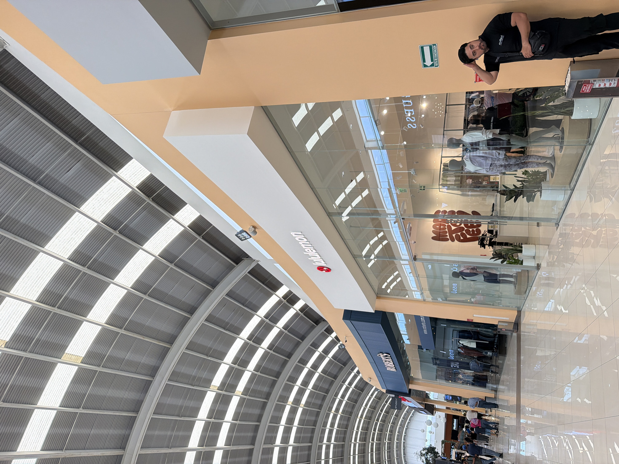 This media file captures the interior of a modern, multi-level shopping mall in San Francisco Ocotlán, Mexico, during the daytime. The perspective is tilted, showing an arched, skylit roof with white structural beams and corrugated metal panels, allowing abundant natural light to illuminate the space.
In the foreground, a man with a beard, wearing a black polo shirt (possibly with a "Quebec" logo), black pants, and a black sling bag, looks directly at the camera with a slight smile or smirk. He is positioned against an orange wall, next to a green emergency exit sign that reads "RUTA DE EVACUACION" (Evacuation Route).
The mall features multiple storefronts with glass facades. Prominently visible is a "Lululemon" store, identified by its red omega symbol logo and text, and a "ROBERTS" store with a dark blue facade. Below "ROBERTS," a "STUDIO" sign is also visible. Inside the "Lululemon" store, mannequins are displayed wearing athletic apparel, alongside potted plants. Reflections on the glass storefronts show glimpses of other parts of the mall and faint text like "www.roberts.com.mx" and "B22."
Further down the concourse, beyond the stores, other shoppers can be seen walking on the brightly tiled floor. The mall's design emphasizes openness and natural light, creating a bright and clean environment for commercial activity.