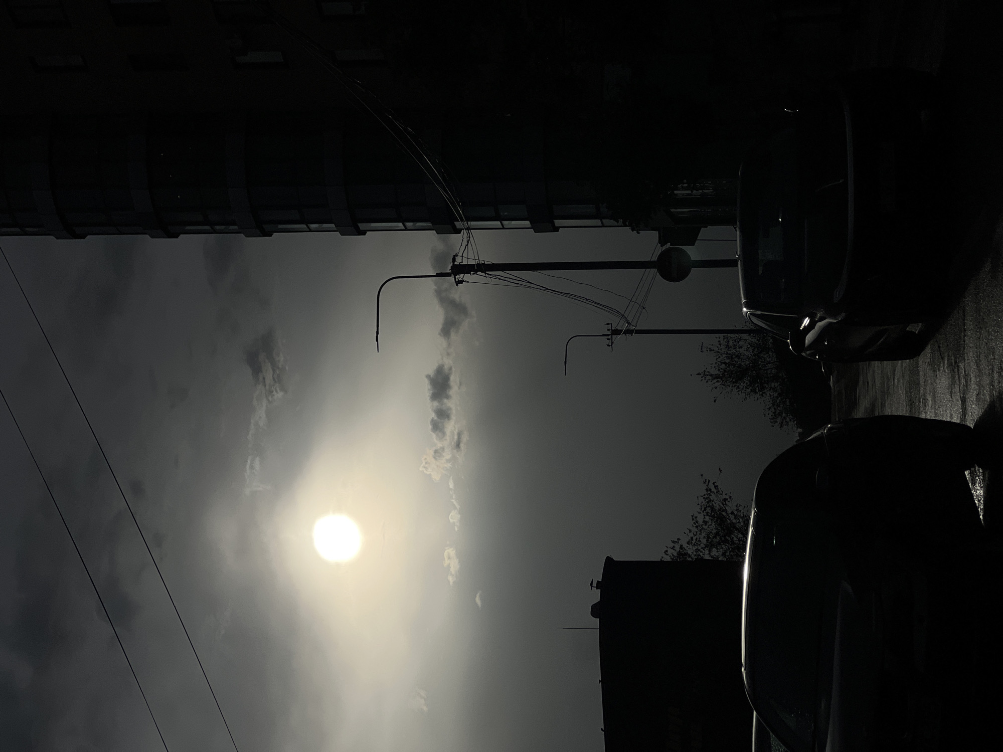 The image depicts a street scene in Yerevan, Armenia, during what appears to be a cloudy day with the sun partially obscured by clouds. The view is oriented upwards, showcasing a cityscape.
On the right, two cars are parked along the street, mostly silhouetted due to the strong contrast of light and shadow. The street surface appears wet, suggesting recent rain.
Overhead, streetlights with wires extend across the sky. Buildings are visible in the background, including a taller structure on the left and another towards the bottom. The sky is overcast, with the sun shining through a break in the clouds.