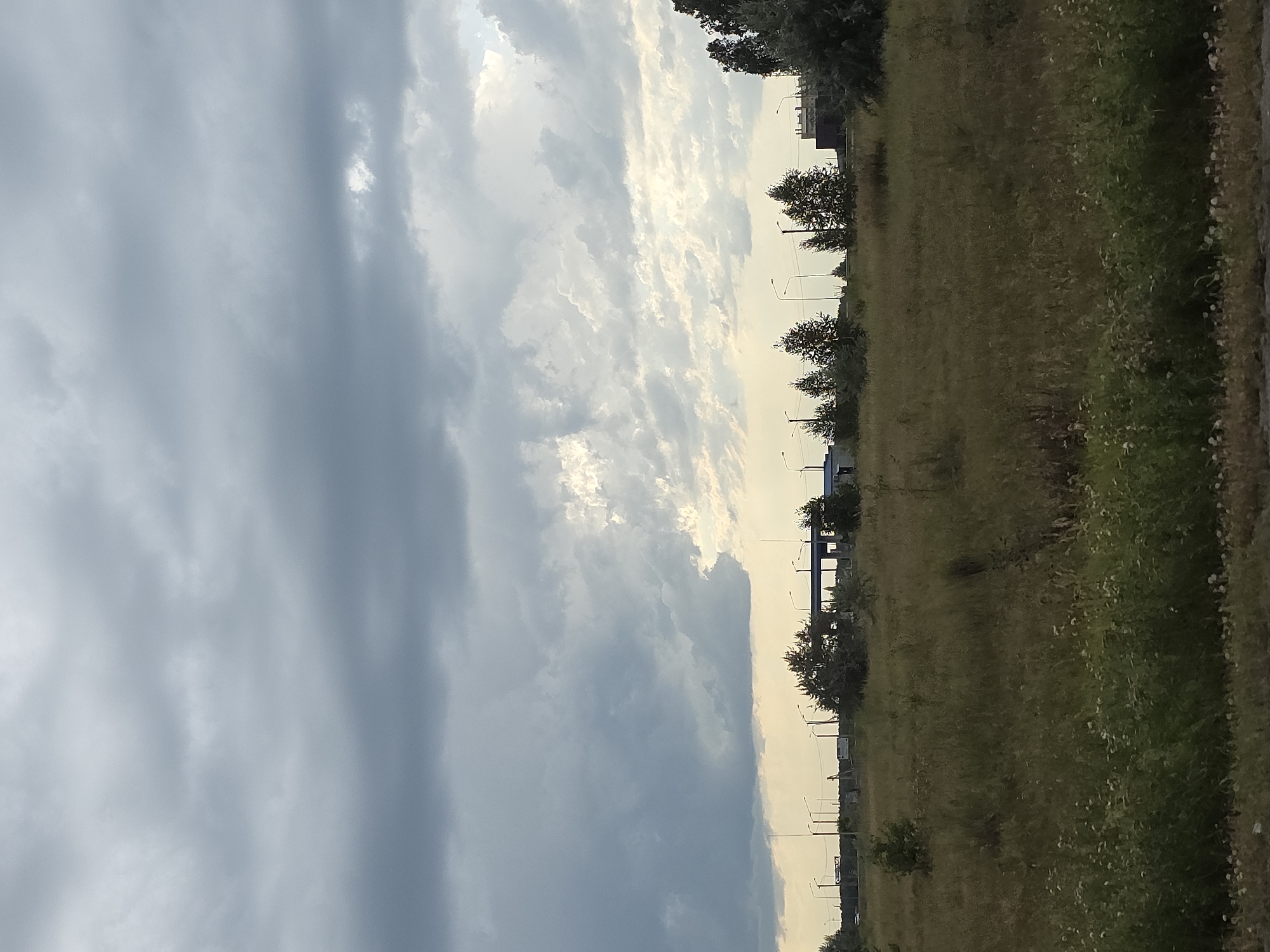 The image depicts a cloudy sky over a rural or suburban landscape. In the distance, there are several utility poles and some low-rise buildings or structures, possibly industrial or commercial. The foreground is dominated by a field of tall grass and wildflowers. The sky is filled with dramatic clouds, with some areas of bright light peeking through, suggesting the possibility of either sunrise or sunset, or a break in the clouds. The overall atmosphere is subdued and somewhat overcast. The location is identified as Karsy, Russia. There are no people or animals visible in the image.