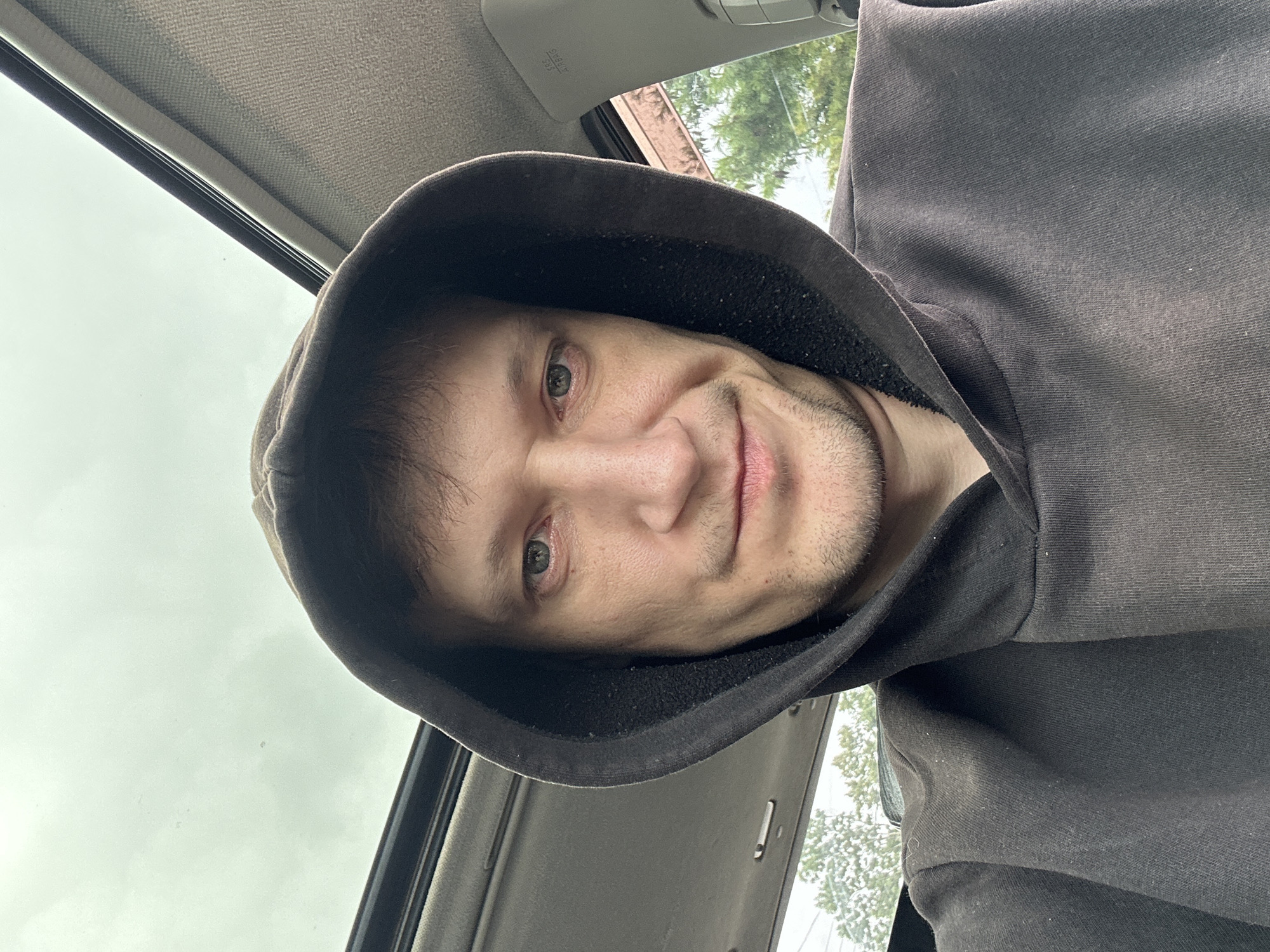 A close-up selfie shows a man wearing a dark hooded sweatshirt inside a car. He is looking directly at the camera with his hood up. The man has light-colored eyes, stubble on his chin and upper lip, and his mouth is slightly curved into a faint smile. The interior of the car is visible, including the headliner and part of the sunroof, which is open, revealing a cloudy sky. Through the open sunroof and the car windows, trees and foliage are visible, suggesting an outdoor setting in a location that appears to be in or around the city of Makiv, Ukraine. The man's facial expression is neutral to slightly amused. The lighting suggests it is daytime, possibly overcast.