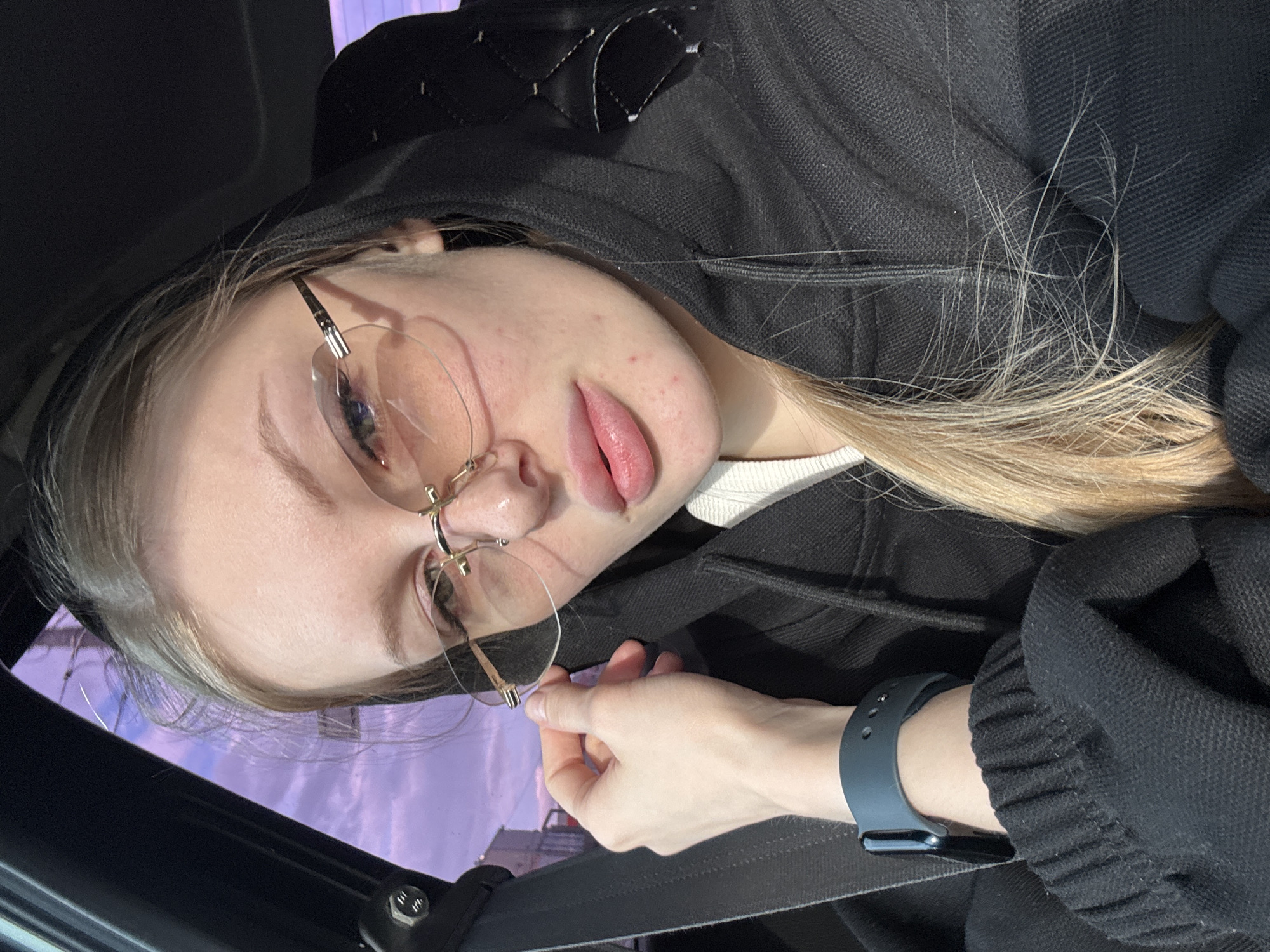 A close-up, rotated portrait shows a young woman wearing glasses and a black hooded sweatshirt, possibly in a car. She has fair skin, blond hair, and is wearing a dark-colored smartwatch on her left wrist. Part of a seatbelt is visible across her chest. The background appears to show an exterior scene with indistinct buildings or structures, and some light purple in the upper left, possibly the sky. Given the prompt indicating the location, the image was potentially taken in Perm, Russia, but without additional information, this cannot be definitively confirmed from the visual details alone.