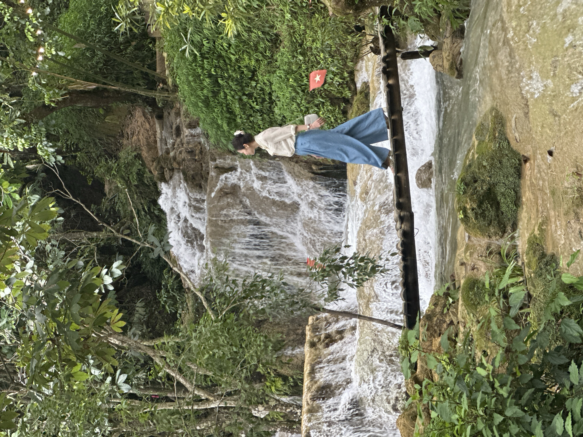 A woman with dark hair pulled back walks across a wooden footbridge over a shallow, fast-flowing river in Cổ Lũng Commune, Vietnam. The woman wears a light-colored, long-sleeved shirt, wide-legged blue pants, and beige shoes. She carries a light-colored bag on her left shoulder. The footbridge is constructed of dark wood planks supported by vertical wooden posts. The riverbed is composed of light-colored rocks and sand, with moss-covered rocks visible along the banks. Behind the river, a multi-tiered waterfall cascades down a rocky slope. The surrounding environment is lush and green, with dense foliage, trees, and bamboo. Two small red flags with white stars are planted on the right bank of the river. The overall scene depicts a natural landscape with a woman enjoying a walk.