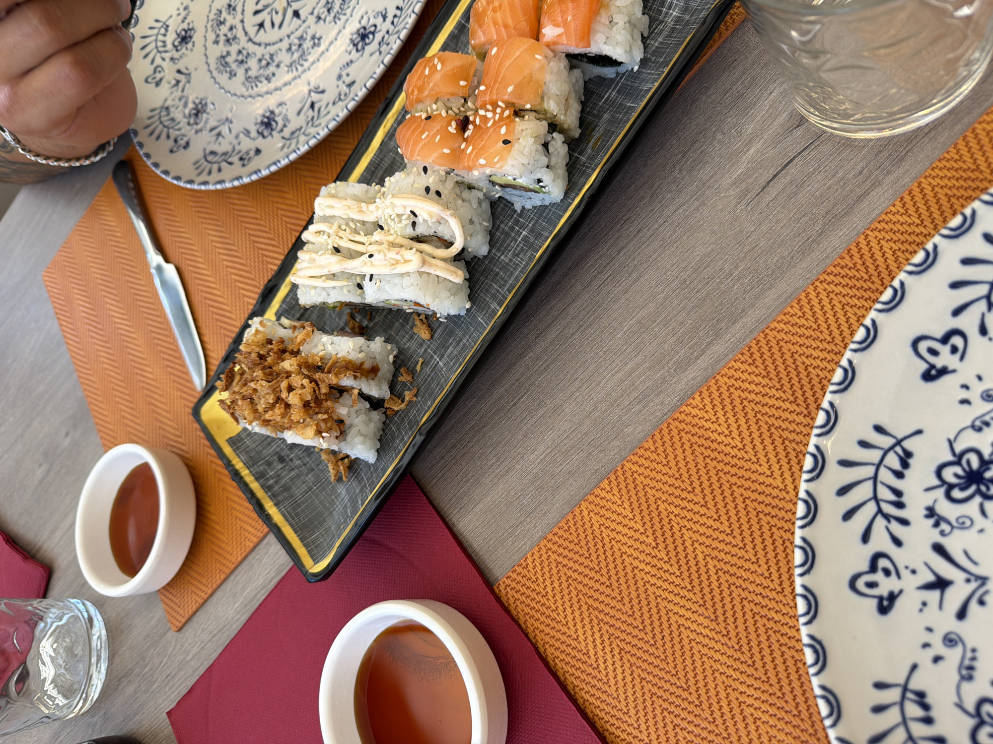 The image displays a close-up, overhead view of a table setting for a meal, likely sushi, in a restaurant in Benidorm, Spain. The table surface appears to be made of a light-colored wood laminate.
On the table, several elements are arranged:
* **Tableware:** A dark rectangular plate with a gold trim holds several pieces of sushi. To the left, two small white ceramic bowls contain a dark reddish-brown liquid, presumably soy sauce or a similar dipping sauce. A white ceramic plate with a blue floral pattern is partially visible in the foreground. A textured orange placemat lies beneath the sushi plate, and a dark red placemat is positioned to the left, beneath one of the soy sauce bowls. A metallic knife is placed on the orange placemat, to the right of the sushi. A clear glass with a textured pattern is visible in the lower right corner, possibly a water glass.
* **Food:** The sushi appears to be various types, some topped with crispy fried onions, others with a creamy sauce and sesame seeds, and some with slices of salmon. The salmon is bright orange and appears fresh.
* **People:** A person's arm and hand, adorned with a silver bracelet, are visible in the upper right quadrant of the image, suggesting they are reaching for or interacting with the table setting. The person's skin tone indicates they are likely of European descent.
* **Environment:** The lighting suggests an indoor setting, possibly during the daytime, with a warm ambient light. The overall scene conveys a sense of dining at a restaurant. The specific location being Benidorm, Spain, is inferred from the context of the request, although no explicit visual cues in the image itself confirm this.