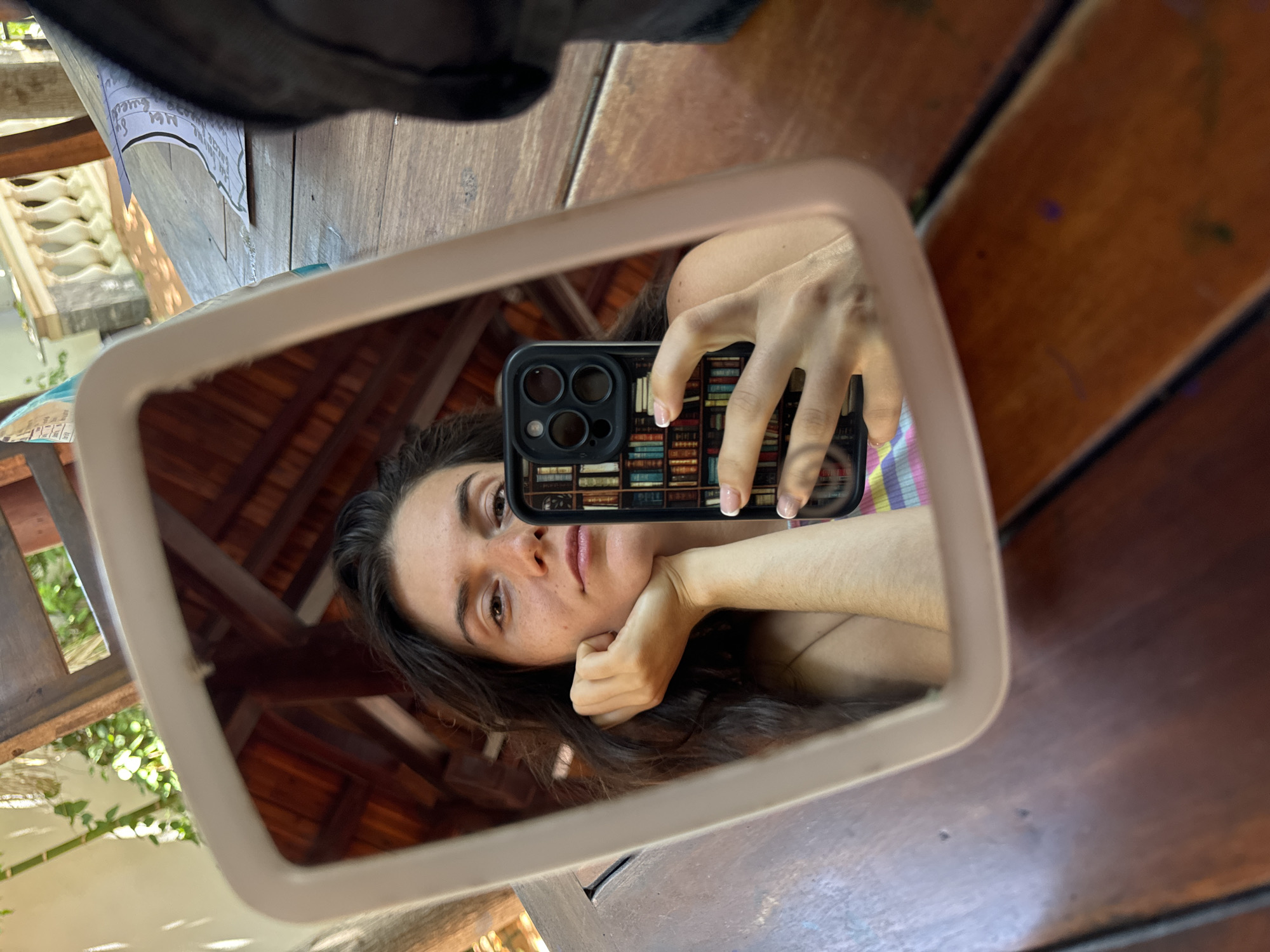 In Mũi Né, Vietnam, a young woman takes a selfie in a mirror. She is leaning her chin on her hand, looking directly at the camera with her hair falling around her face. The mirror reflects the underside of a wooden roof structure and lush greenery, suggesting an outdoor setting. She is holding a smartphone with a phone case designed to look like a bookshelf. Her outfit appears to be a colorful striped top. The background shows a wooden table and some papers. The overall scene is informal and casual.