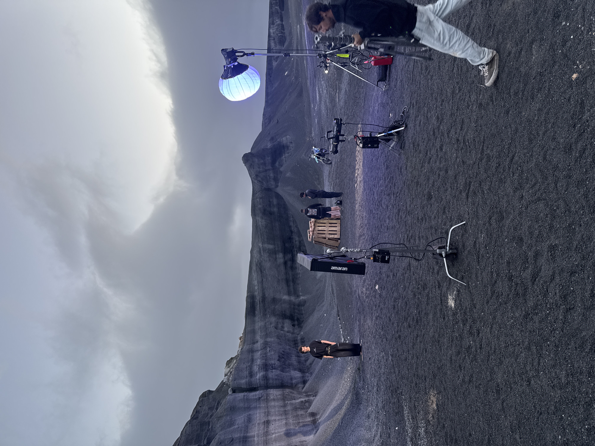 The image shows a film set located in an area of volcanic ash, seemingly in Teguise, Spain. The landscape includes a layered rock formation on the left side of the frame. The sky is overcast.
Several individuals are present. A man wearing a black jacket and light-colored pants is partially visible at the top of the frame, operating some equipment. Near the center, a group of people stand near wooden pallets. In the foreground, a man in black clothing stands facing the camera.
There are several pieces of film equipment on set including a large spherical light suspended on a rig, a lighting apparatus with an "amaran" logo, and a motorcycle. Cables and stands are visible across the landscape.