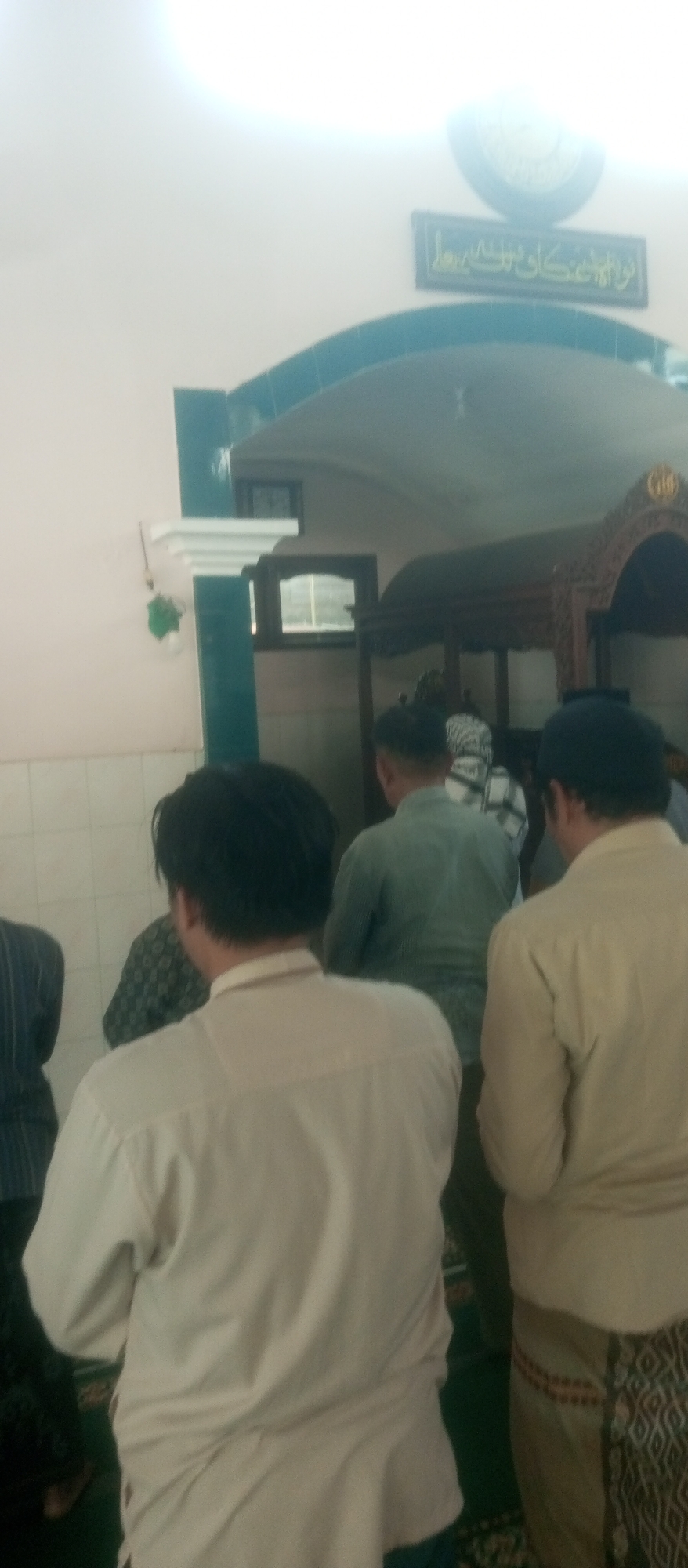 The image shows the rear view of several men inside a building, likely a mosque, given the architectural elements. In the foreground, a man in a light-colored long-sleeved shirt faces away from the camera. To his left, partially obscured, is another individual. To the right of the foreground man, another man wears a similar light-colored shirt and a patterned lower garment. Behind them, a man in a green long-sleeved shirt is visible. Further back, other individuals can be seen, along with architectural features such as an ornate wooden structure, a tiled archway with turquoise trim, and a sign with Arabic calligraphy. The lighting suggests an indoor setting with some natural light. The image's context points to a religious gathering or prayer service.