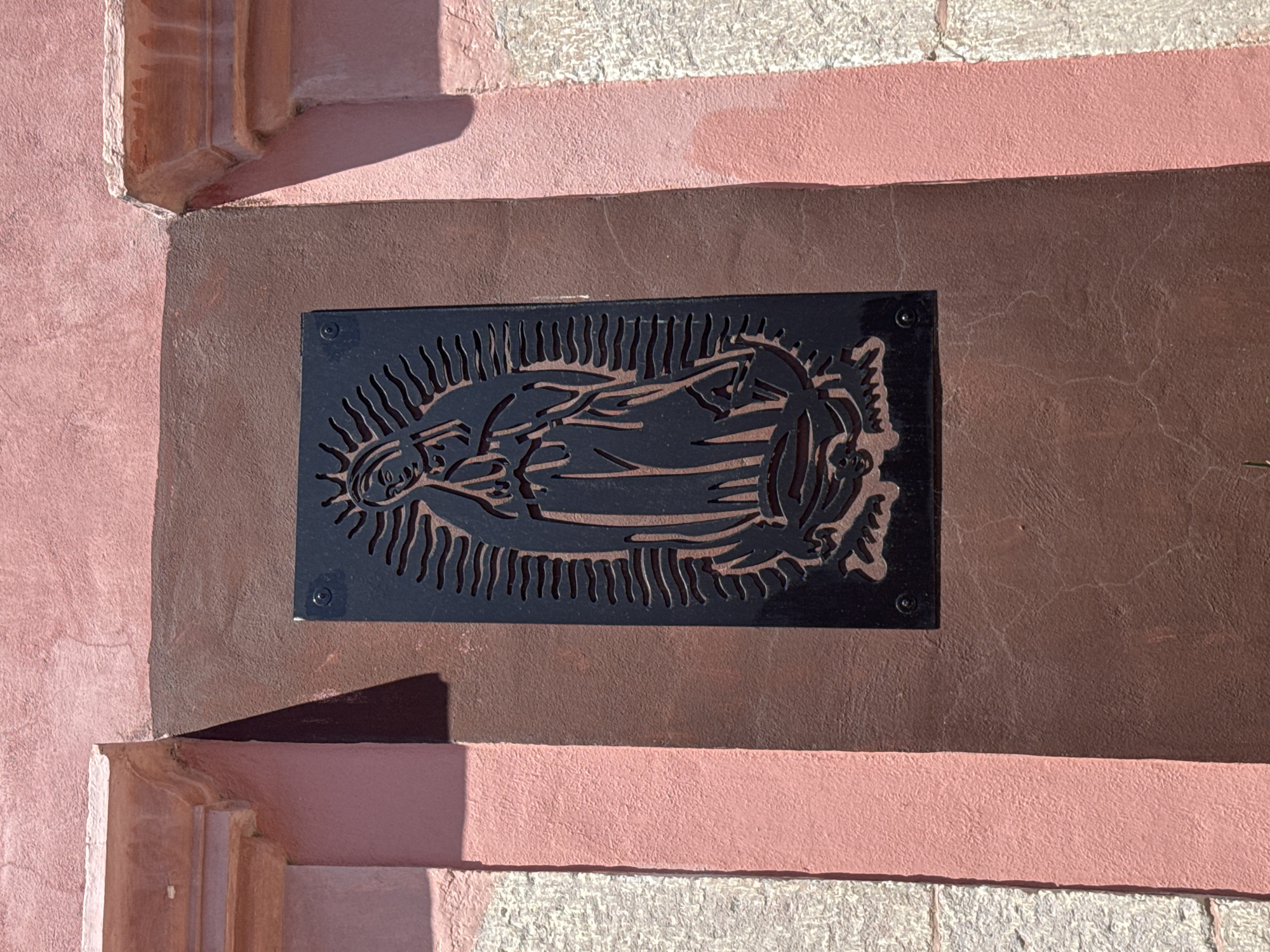 A close-up shot reveals a black metal plaque depicting Our Lady of Guadalupe, mounted on a textured brown wall with traces of pink paint along the edges. The plaque is secured with screws at each corner. The surrounding architecture suggests an exterior wall, possibly in Bernal, Mexico.
