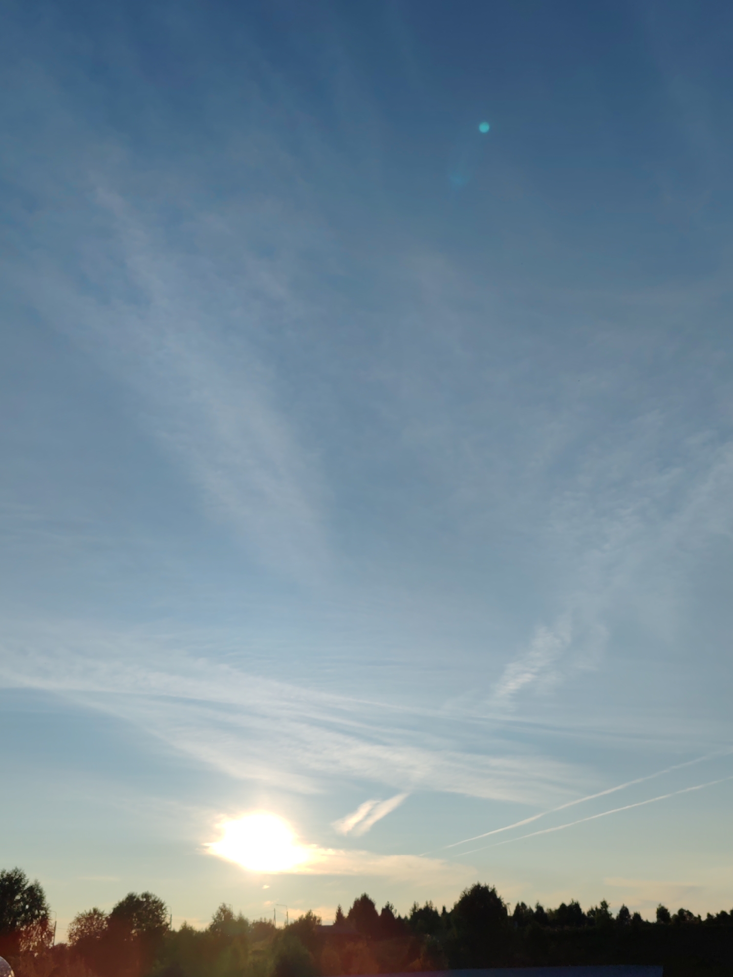 The image depicts a wide-angle view of a sunset over a treeline, with a bright sun low on the horizon. The sky is a gradient of light blue to a pale yellow near the sun, with wispy white clouds scattered across it. Two distinct contrails, likely from aircraft, are visible. The foreground is dominated by a dense line of dark green trees and foliage, suggesting a natural or semi-rural setting. No people or specific urban structures are clearly identifiable. The scene appears to be taken from an elevated or open position looking towards the west. The specific location in Чухлэм, Russia, is not visually indicated within the image itself.