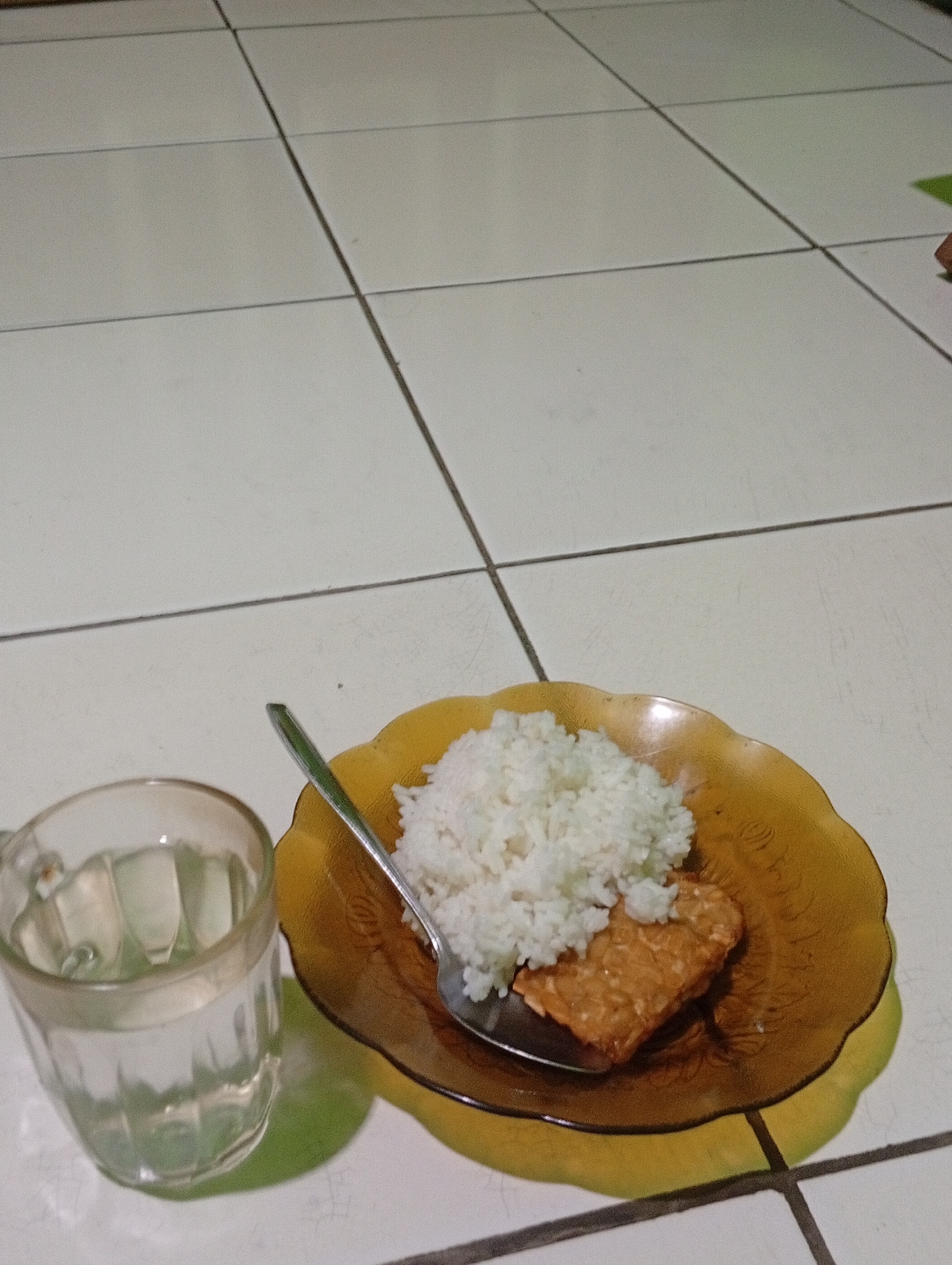 A glass of water and a plate of food sit on a tiled floor. The glass is clear and filled with water, with ripples visible on its surface. The plate is a translucent amber color, with a scalloped edge. It holds a portion of white rice and a piece of fried tempeh. A silver spoon rests on the plate, partially submerged in the rice. The floor is made of white square tiles with dark grout lines. The scene appears to be indoors, possibly in a kitchen or dining area in Nyalindung, Indonesia. The lighting is dim, casting shadows on the floor. There are no people visible in the image.