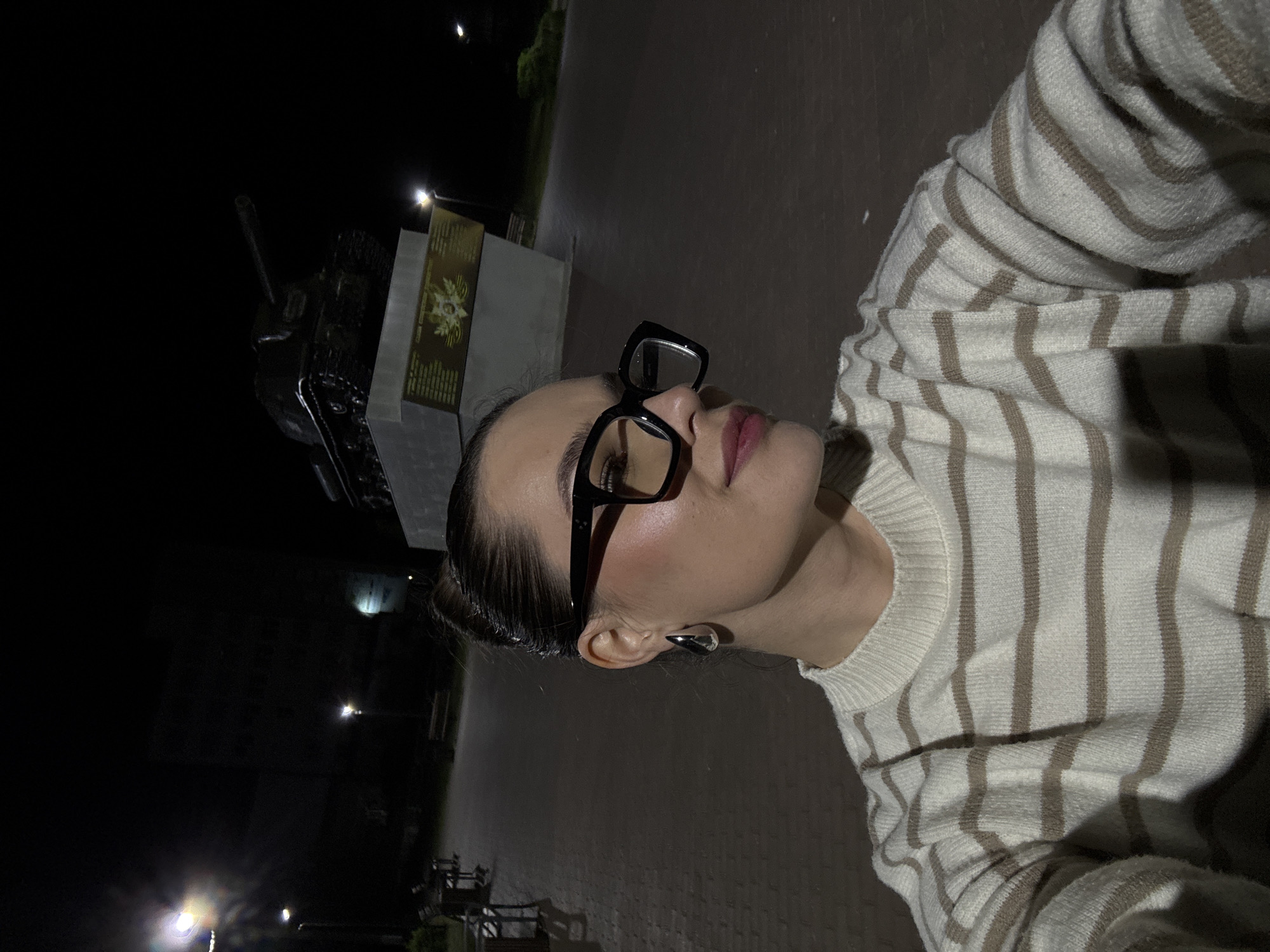 A young woman with dark hair pulled back in a bun, wearing a cream-colored sweater with horizontal brown stripes and large black-rimmed glasses, is taking a selfie at night. She is looking to her left with her head slightly tilted. Her left ear is adorned with a large, teardrop-shaped silver earring. In the background, a monument featuring a tank on a raised platform is visible, illuminated by artificial lighting. The surrounding area appears to be a public space with paved walkways and benches. This scene is located in the city of Chaikovsky, Russia, specifically at the memorial complex dedicated to the Great Patriotic War, which houses a tank monument and commemorates fallen soldiers.