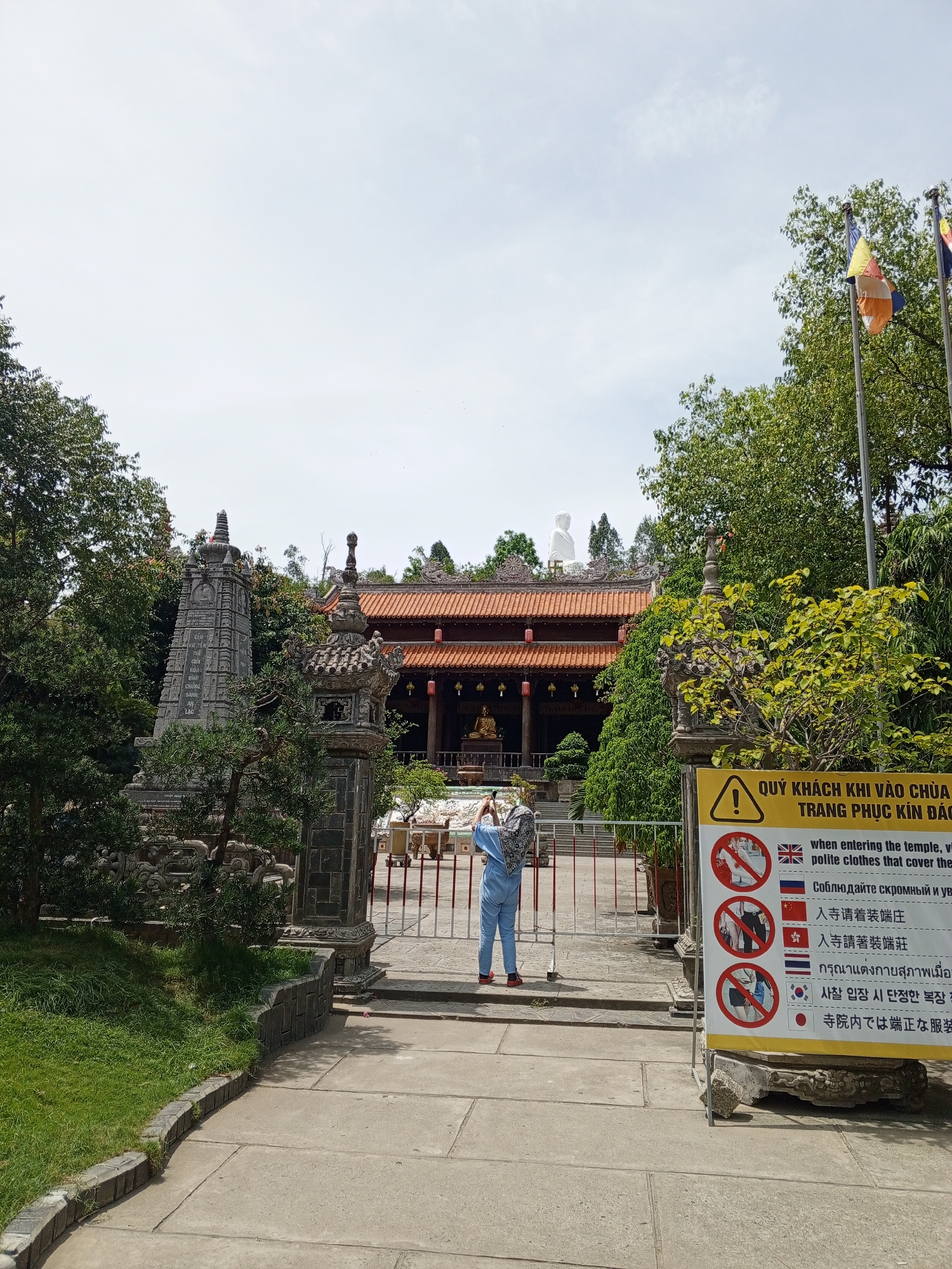 The image depicts the entrance to a temple complex in Tay Nha Trang Ward, Vietnam. In the foreground, a paved walkway curves around a well-maintained grassy area. A person in a blue outfit with a patterned scarf is standing near a gate, holding a camera and appearing to take a photograph. To the right of the gate is a large yellow sign with several multilingual instructions and warning symbols, including images prohibiting immodest clothing. The sign is supported by a stone base. Beyond the gate, a traditional temple structure with an orange tiled roof is visible, flanked by trees and stone pillars. A large white Buddha statue stands atop the temple roof in the background. Flags are also visible on poles to the right, partially obscured by trees. The scene suggests a cultural and religious site open to visitors, with guidelines in place for appropriate attire.
