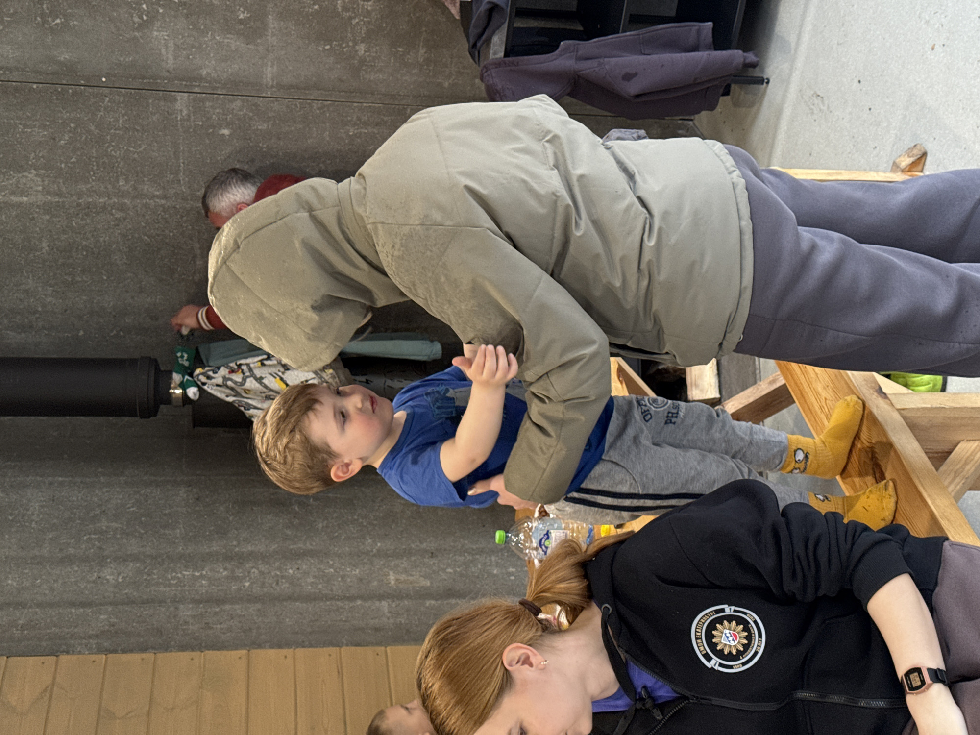 The image, captured in an indoor setting in Yekaterinburg, Russia, depicts several individuals engaged in various activities. In the central focus, a man wearing a light grey hooded jacket and dark grey sweatpants is leaning forward, interacting with a young boy. The man's body is positioned above the child, suggesting he is offering assistance or playing.
The young boy, dressed in a blue t-shirt and grey sweatpants adorned with "OFFICIAL PHS" text, is seated on a low, rustic wooden structure. He wears distinctive yellow socks and looks upwards towards the man with an engaged expression. Adjacent to the boy on the wooden structure is a plastic water bottle, identifiable by its "BON AQUA STILL" label and green cap.
In the foreground, partially visible, is a woman with reddish-brown hair tied in a ponytail. She is wearing a black zip-up hoodie featuring a circular emblem on the left side, which clearly displays the words "PHILIPPINE SPORTS COMMISSION" and the year "1990". A digital watch is visible on her left wrist, and her gaze is directed downwards.
In the background, another adult with grey hair, wearing a red hoodie, is partially visible. The overall setting suggests a casual, possibly communal, indoor space with grey concrete walls and wooden paneling or furniture elements. The lighting appears to be ambient indoor light.