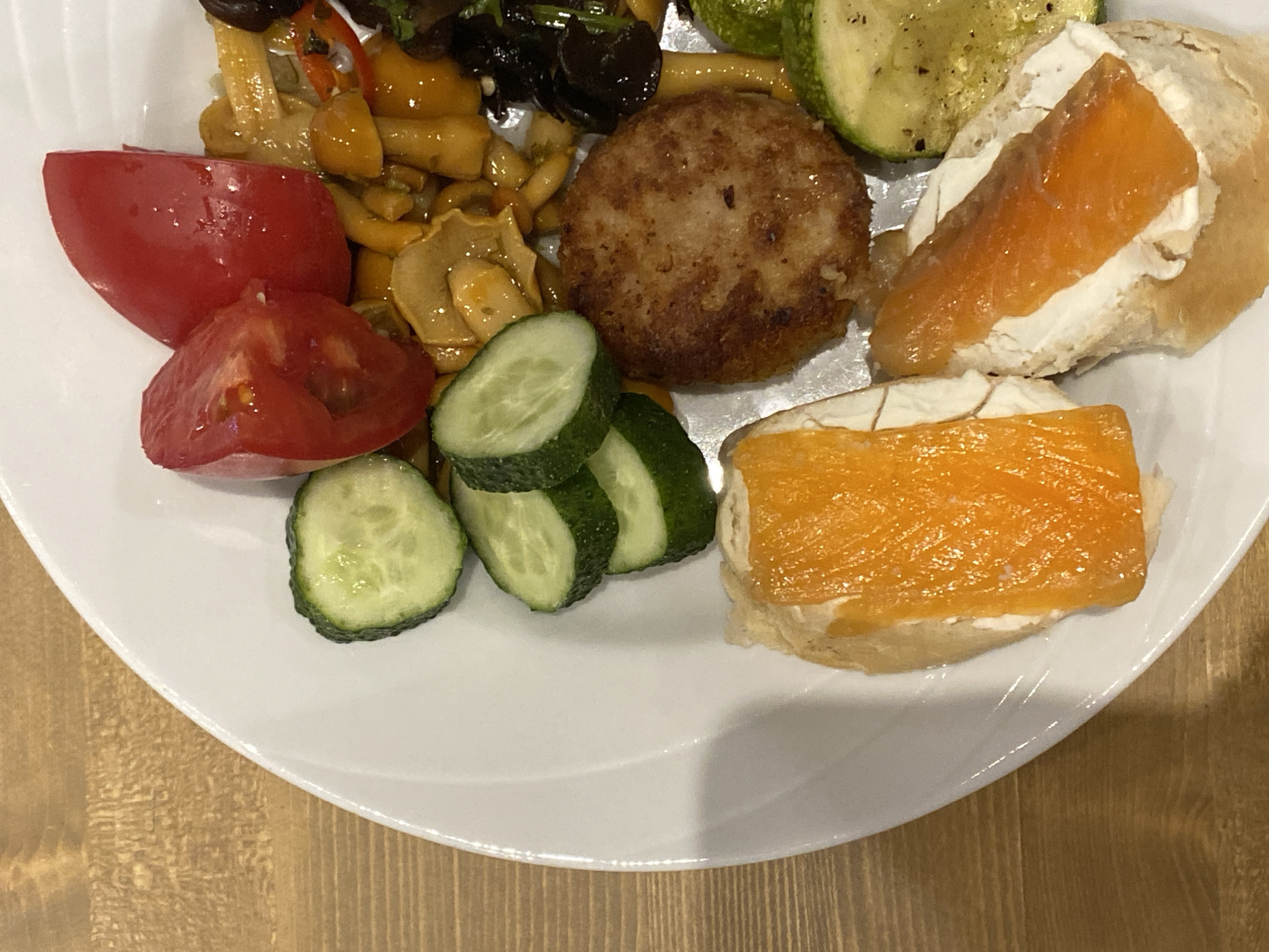 A white plate holds a meal consisting of a single hamburger patty, slices of cucumber, chopped tomatoes, and two pieces of bread topped with cream cheese and smoked salmon. The plate rests on a wooden surface, suggesting an indoor setting. The lighting is somewhat dim, possibly indicating an evening meal. The food appears to be ready to be eaten, and there are no people visible in the frame. The overall presentation is casual, implying a home-cooked meal or a relaxed dining experience. The location context of Krasnoflotskoe, Russia, does not provide specific visual cues within the image. There is no visible text in the image.