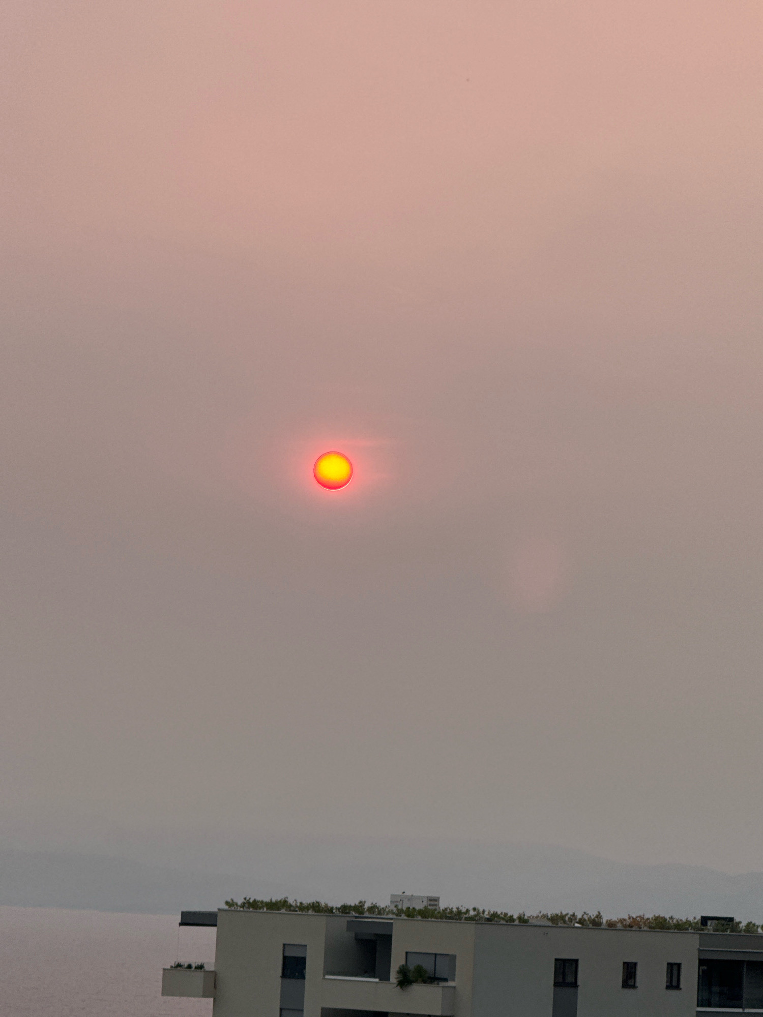 The image captures a hazy sunset over the city of Makarska, Croatia. The sun, a bright orange-red disc, is partially obscured by atmospheric particles, casting a diffused glow across the sky, which transitions from a pale pink near the top to a muted grey towards the horizon. In the foreground, modern buildings with flat roofs and balconies are visible. Some buildings feature rooftop greenery, suggesting a contemporary architectural style. The scene is dominated by the sky and the muted colors of the environment, likely due to smoke or dust in the air. The water is barely discernible as a pale band along the bottom left, merging with the hazy sky and landforms in the distance. There are no people or animals visible in the frame.